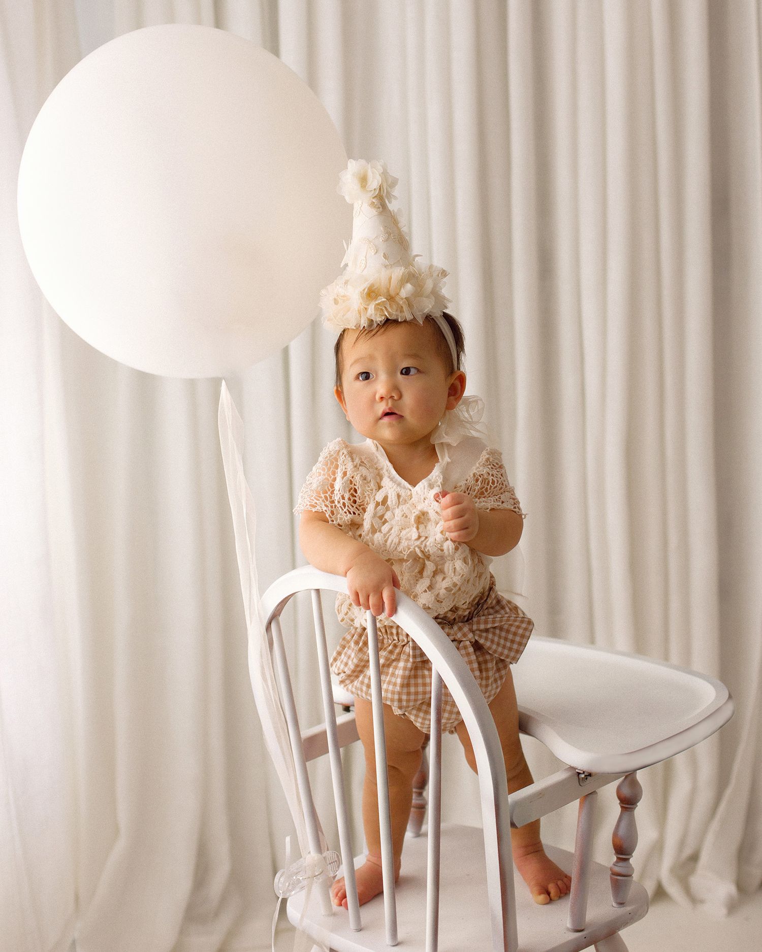 Toddler in a cream floral romper and party hat stands on a white wooden chair beside a large white balloon against soft white curtains.