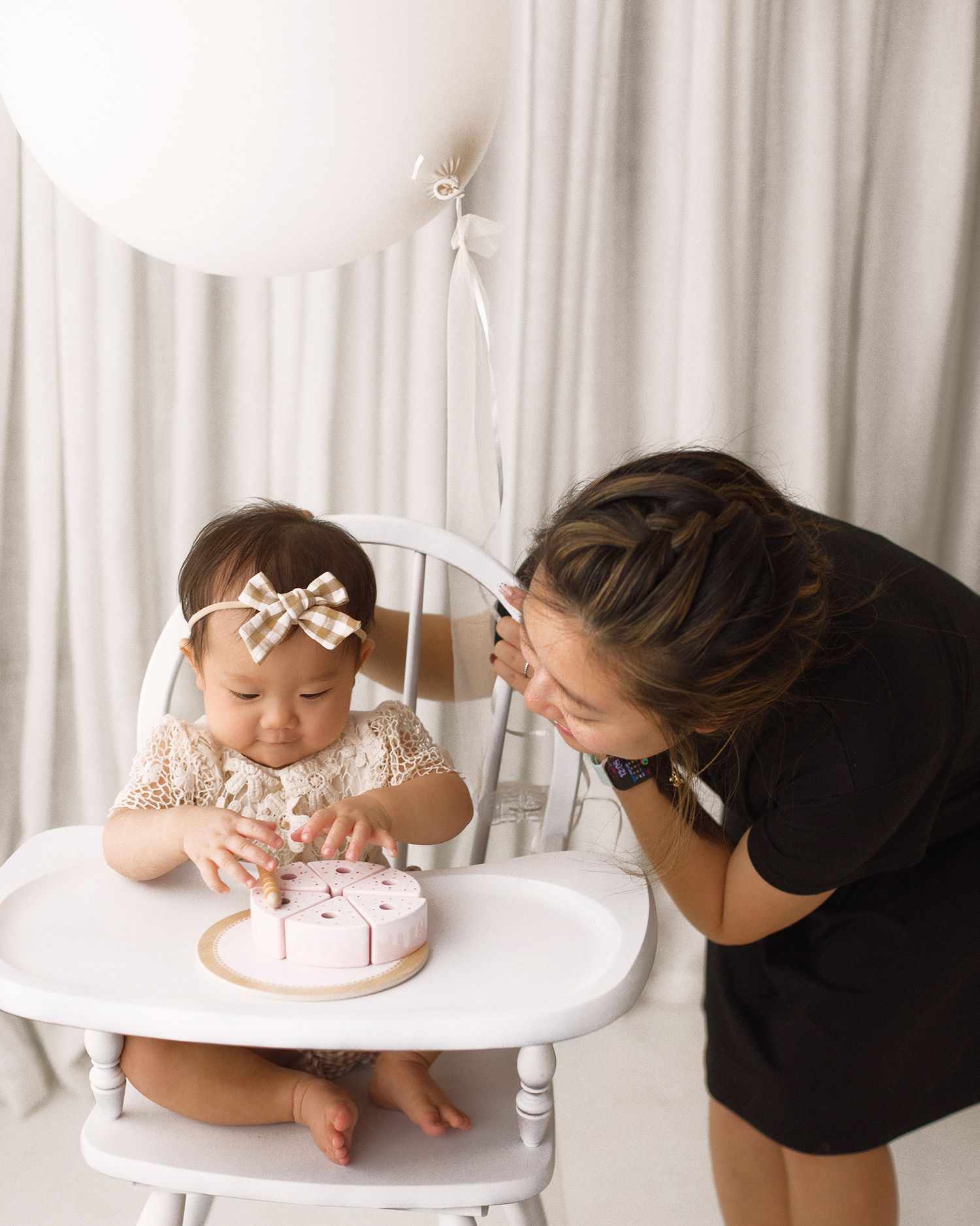 Baby girl in a bow headband sits in a white high chair touching a small pink cake while a smiling woman leans in beside a balloon.