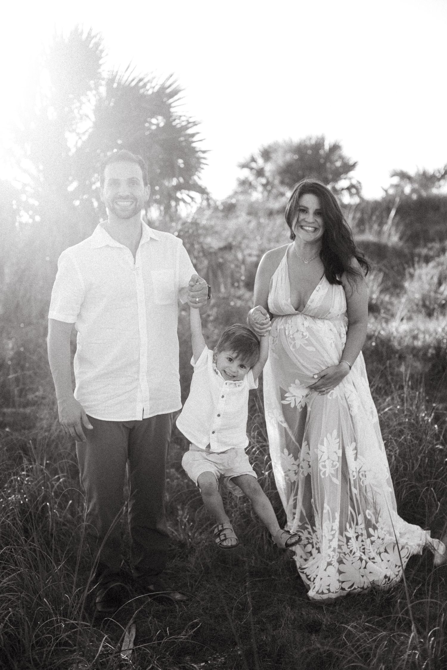 Black and white maternity photo of a happy family with young son in a sunlit field.