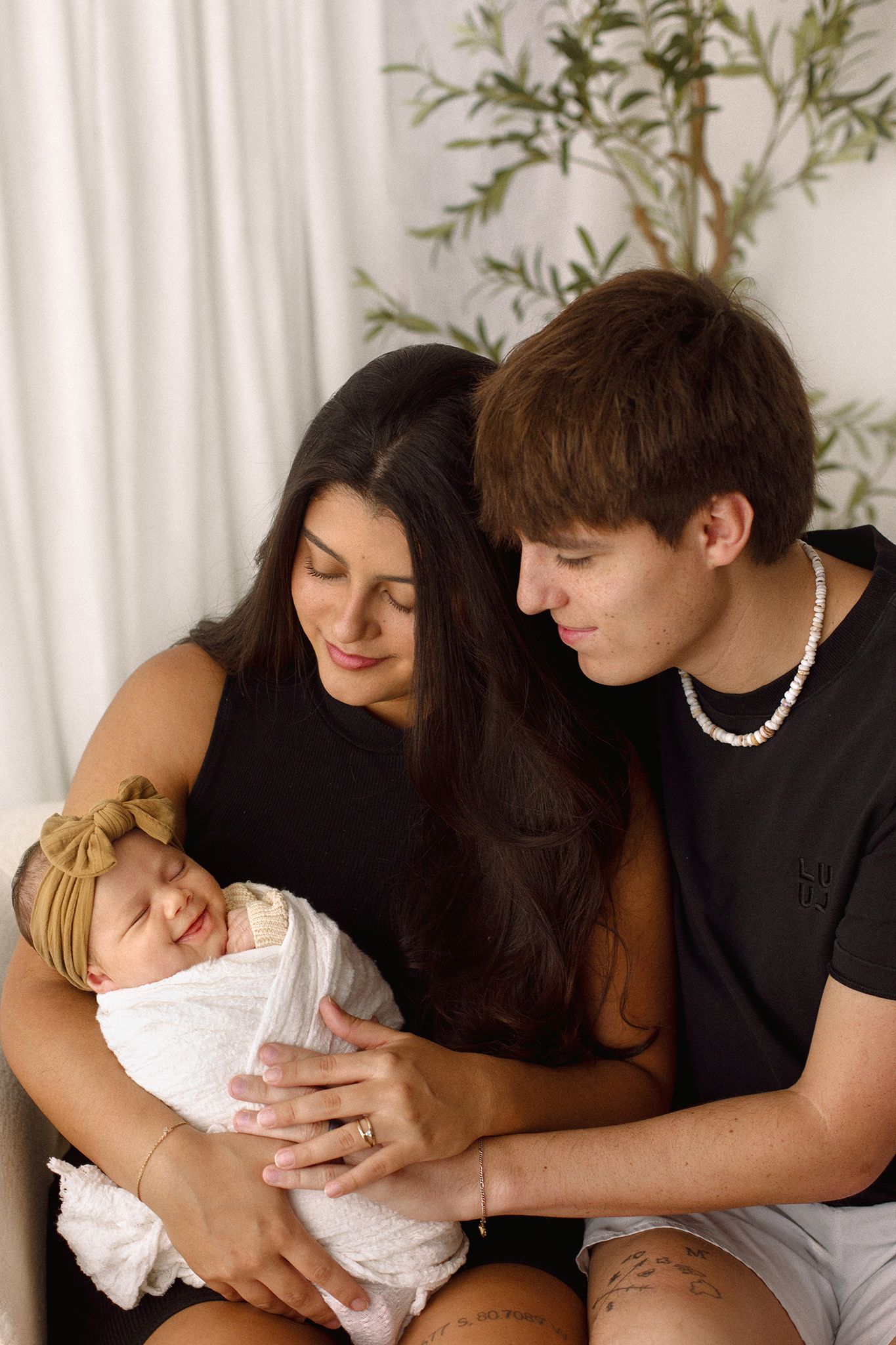 A young couple in black outfits lovingly cradle their sleeping newborn baby wrapped in white cloth with a bow headband.