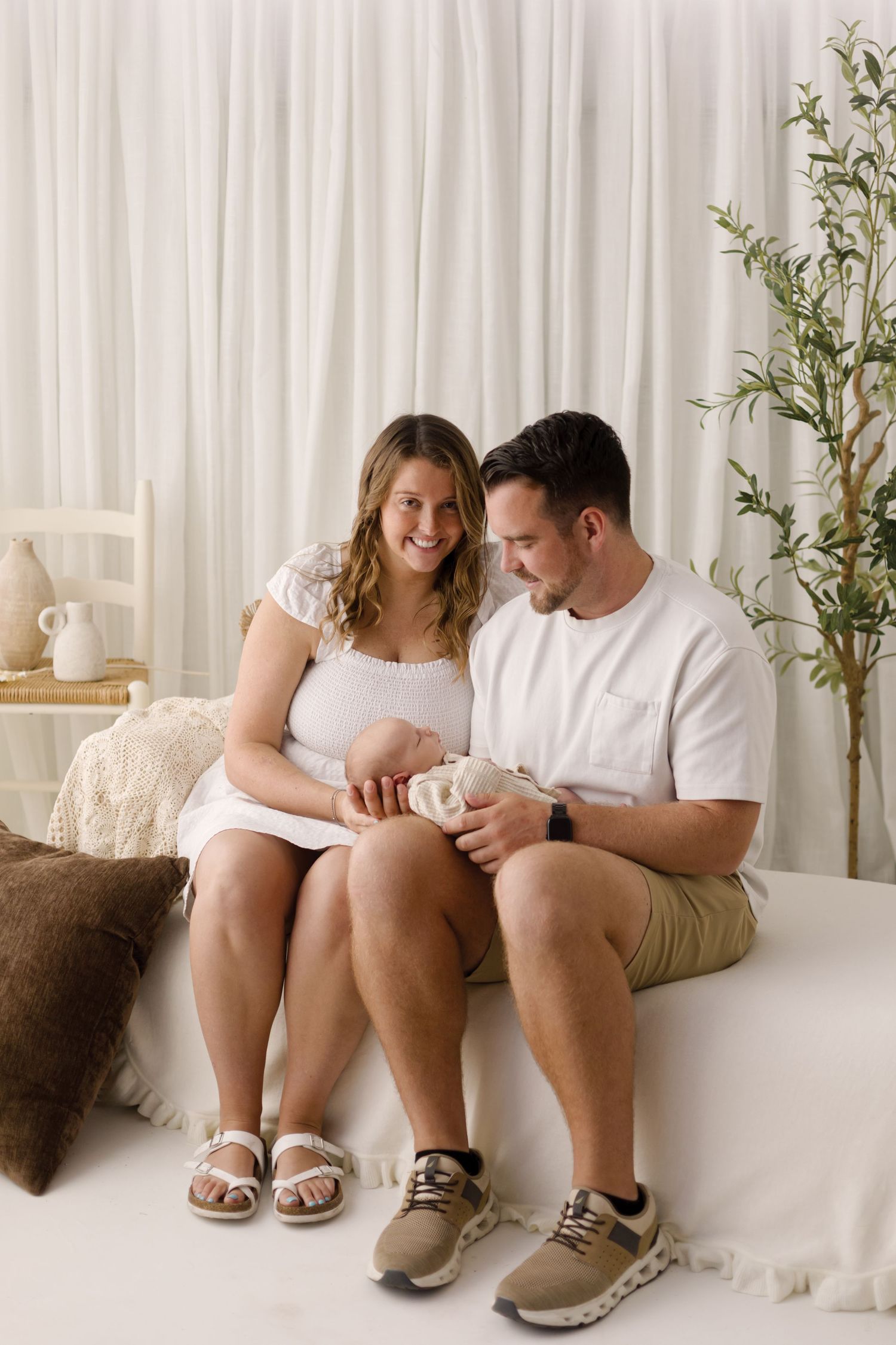Smiling young couple sitting on a bed holding their newborn baby in a bright white room.