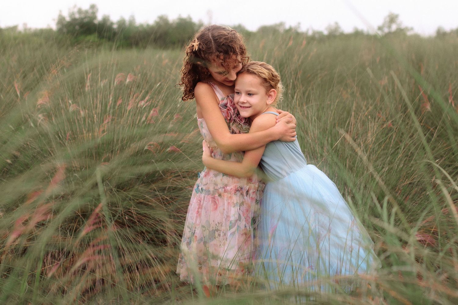 Two young girls in pastel dresses share a warm hug in a tall green grass field on a soft, overcast day.
