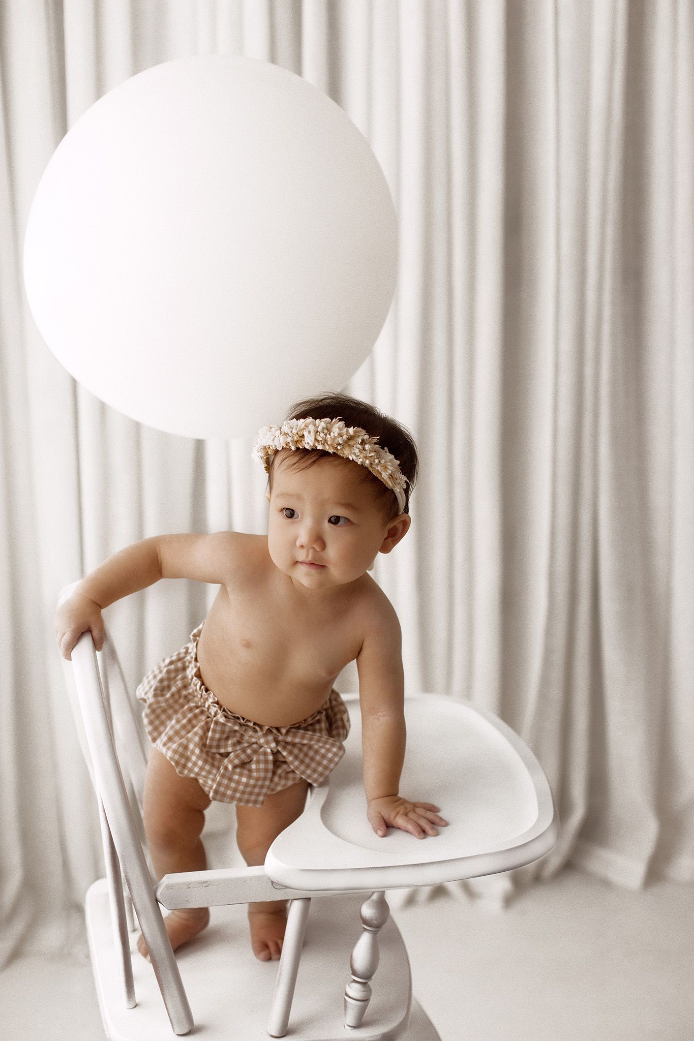 Baby in a gingham bloomer and floral headband stands on a white wooden high chair beside a large white balloon against sheer white curtains.