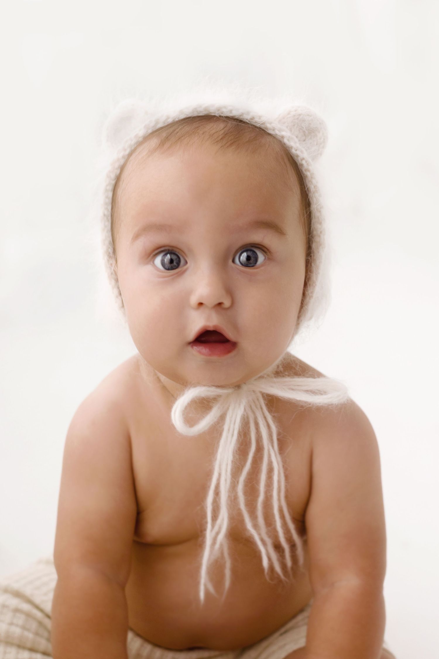 Adorable baby wearing a white knit bear ear bonnet with bow tie strings, looking surprised with wide blue eyes.