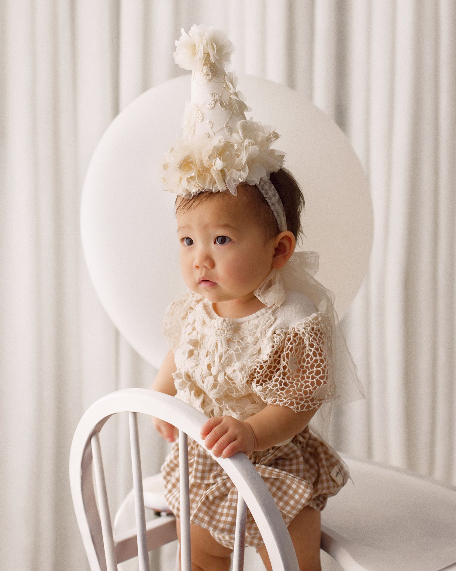Baby in a cream lace romper and floral party hat stands on a white wooden chair against a soft white backdrop.
