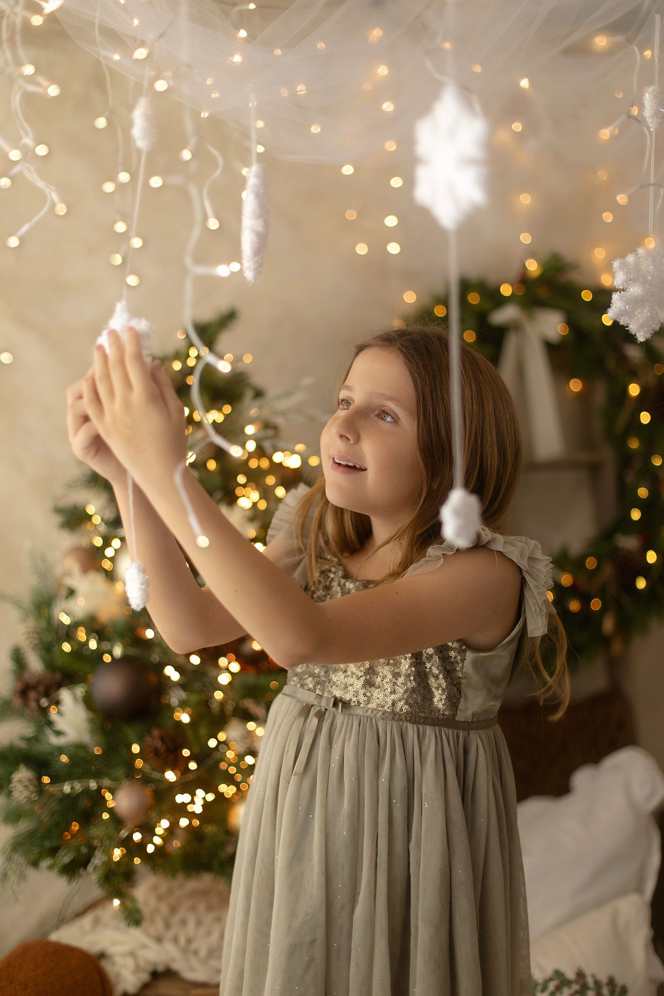Young girl in elegant gray dress decorating Christmas tree with fairy lights and snowflake ornaments in festive room.