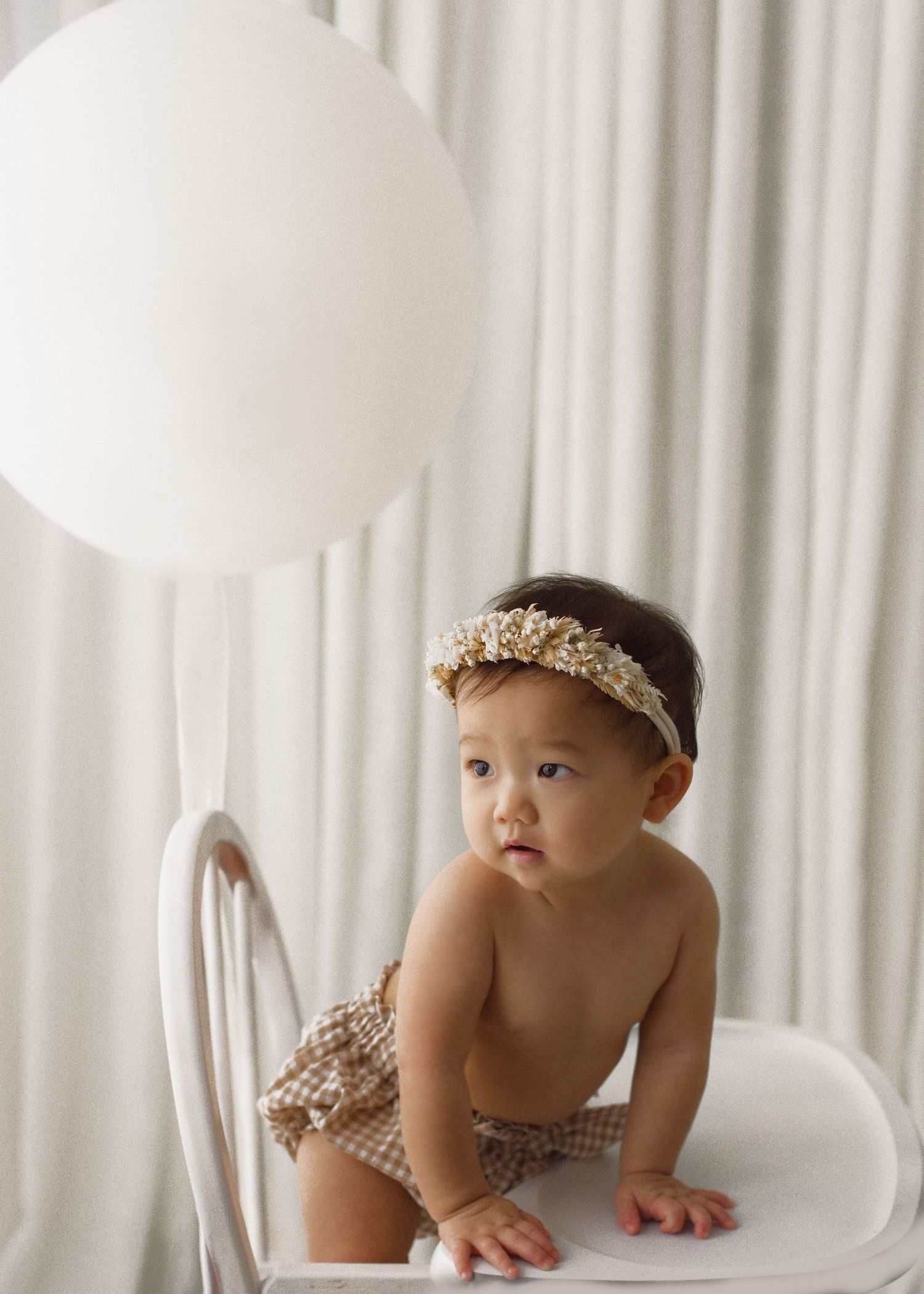 Baby in checkered bloomers and floral headband sits on white chair near large white balloon.