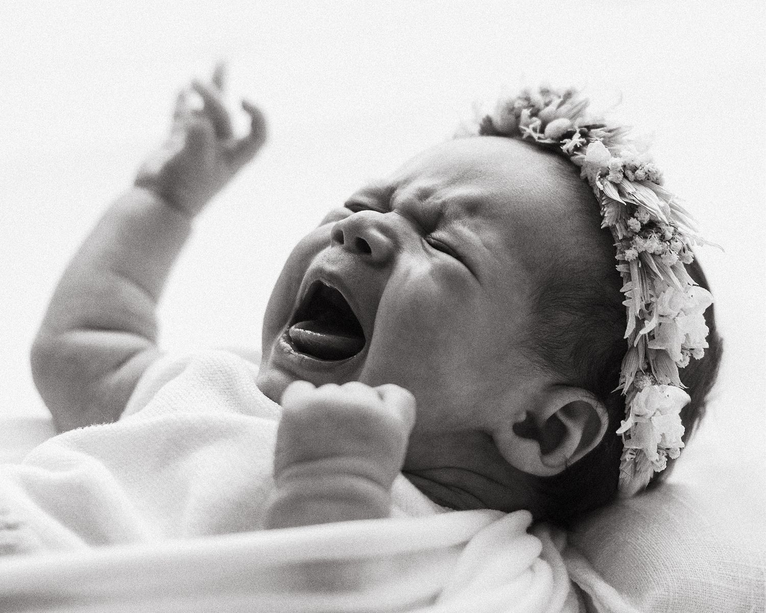 Black and white photo of a crying newborn baby girl wearing a floral headband, arms raised.