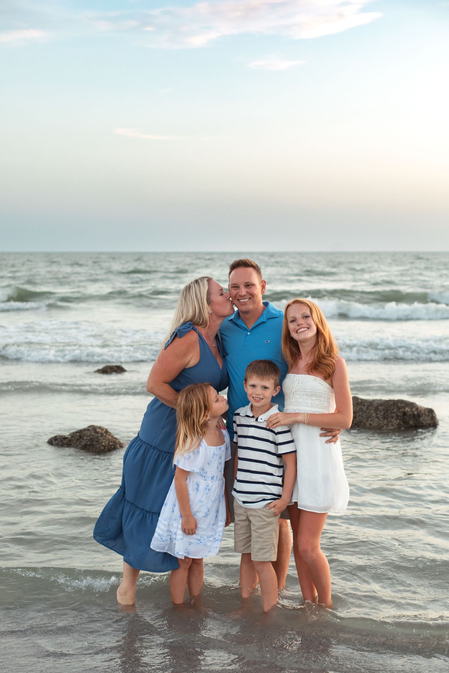 Family of five posing together on a beach at sunset, with waves and rocks in the background.