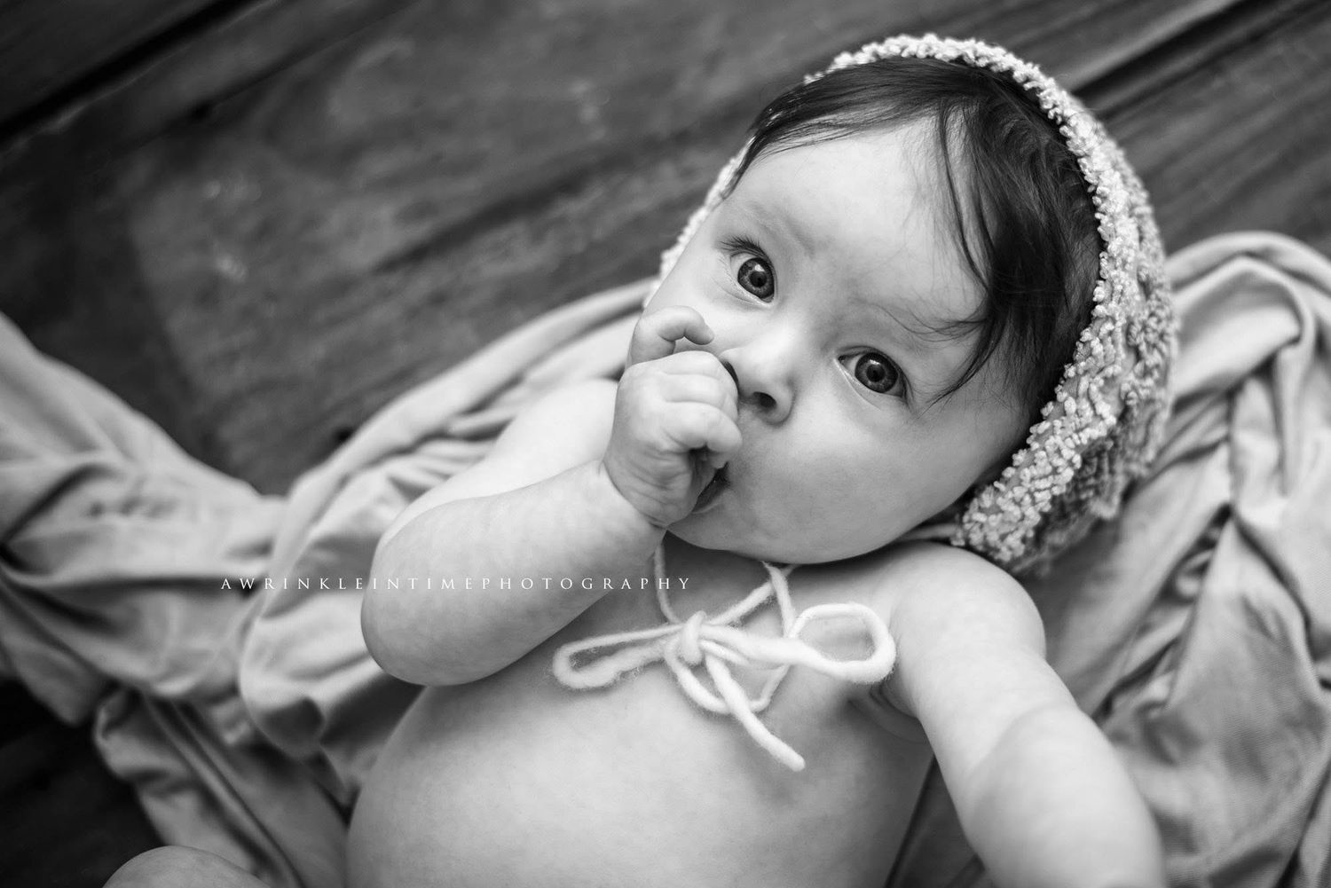 Black and white photo of an adorable baby wearing a floral bonnet, sucking fingers while lying on a soft blanket.