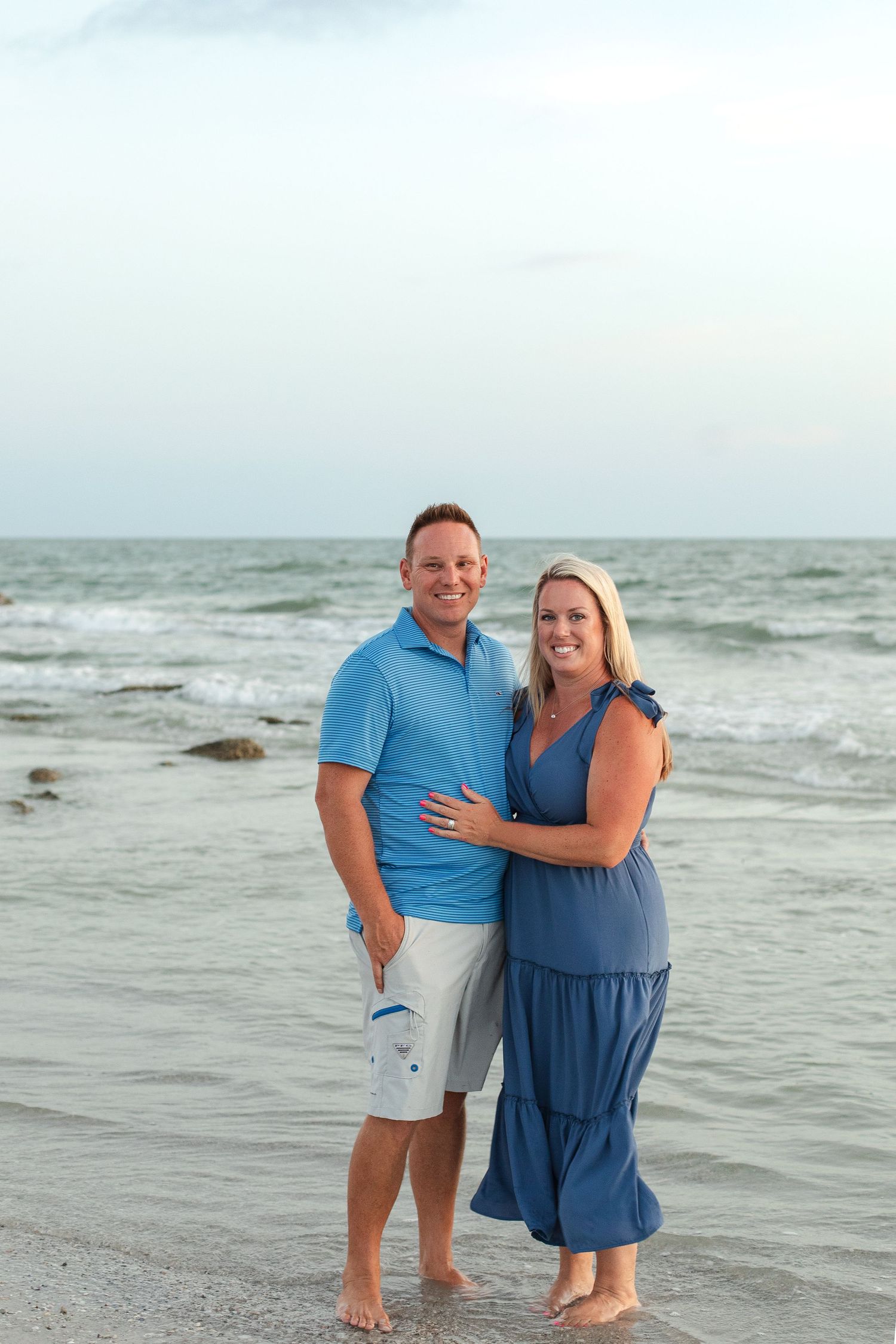 Smiling couple in blue outfits embracing on a sandy beach with ocean waves in the background.