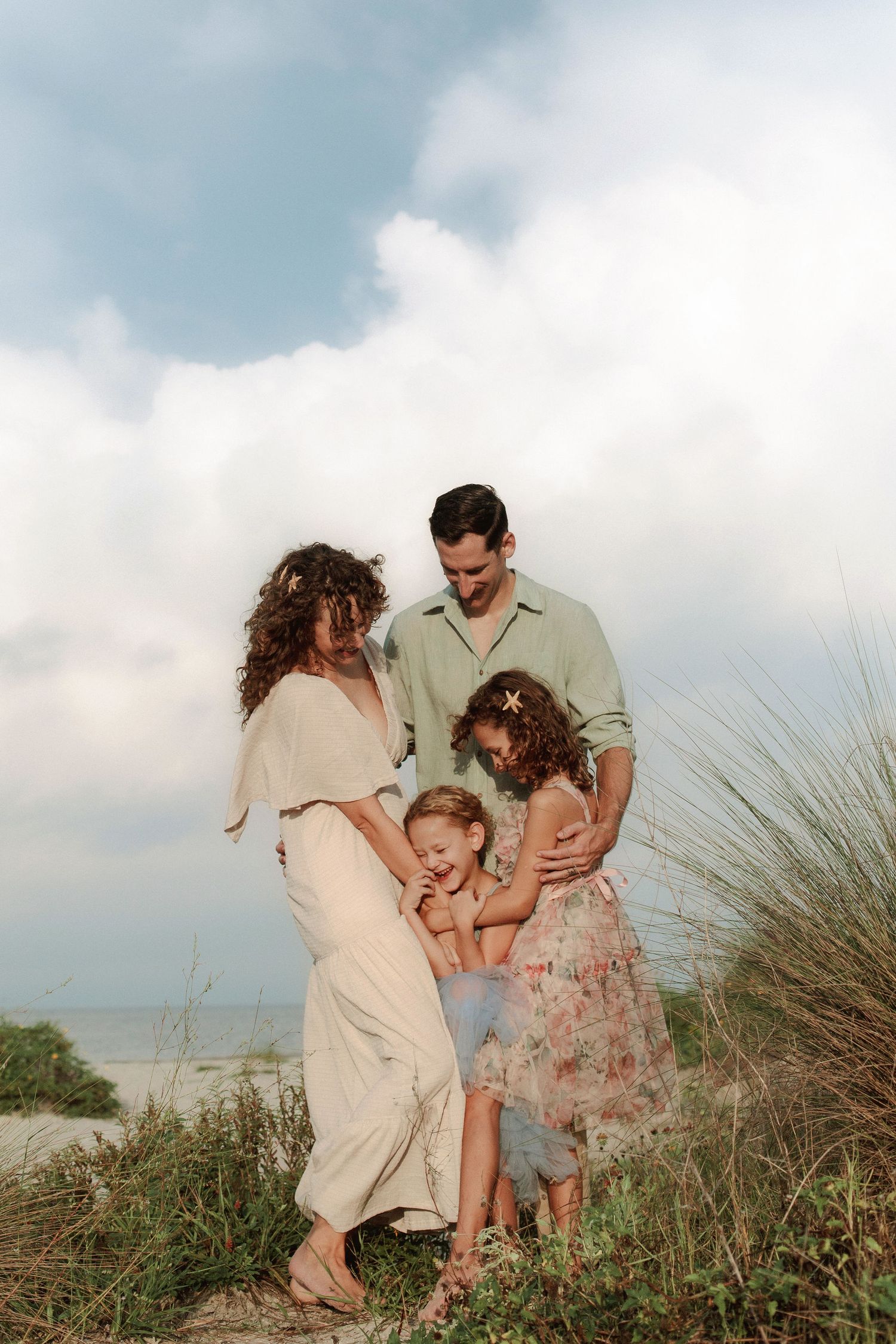 Family of four in light summer outfits sharing a warm embrace on a grassy beach dune under a soft, cloudy sky.