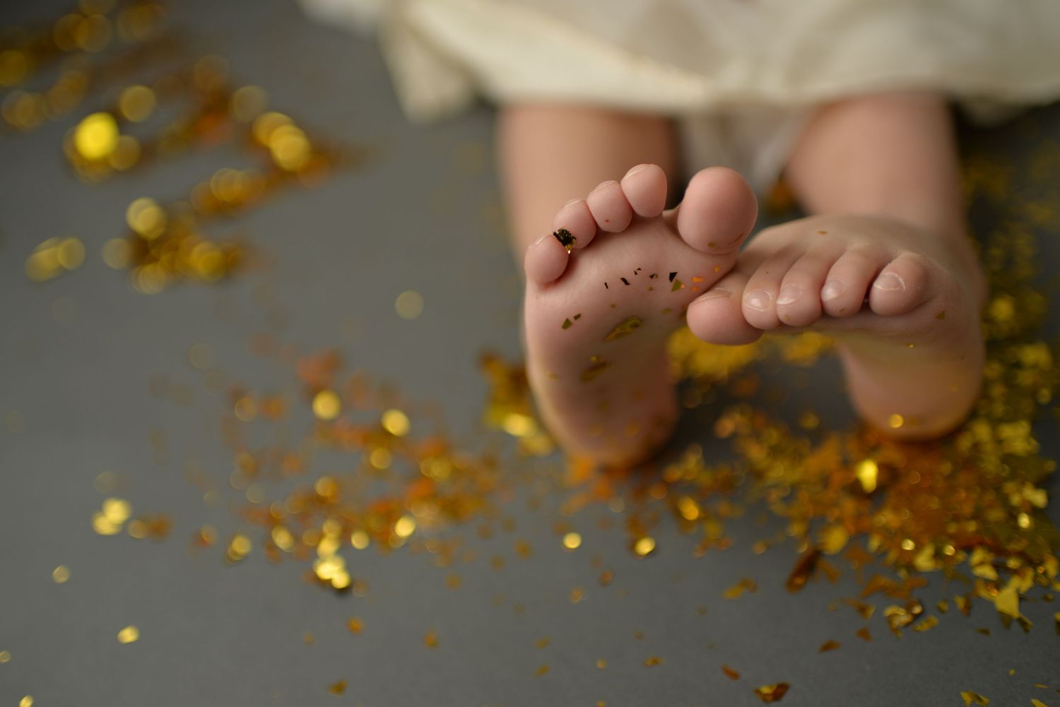 Close-up of tiny baby feet surrounded by golden glitter confetti on a gray background.