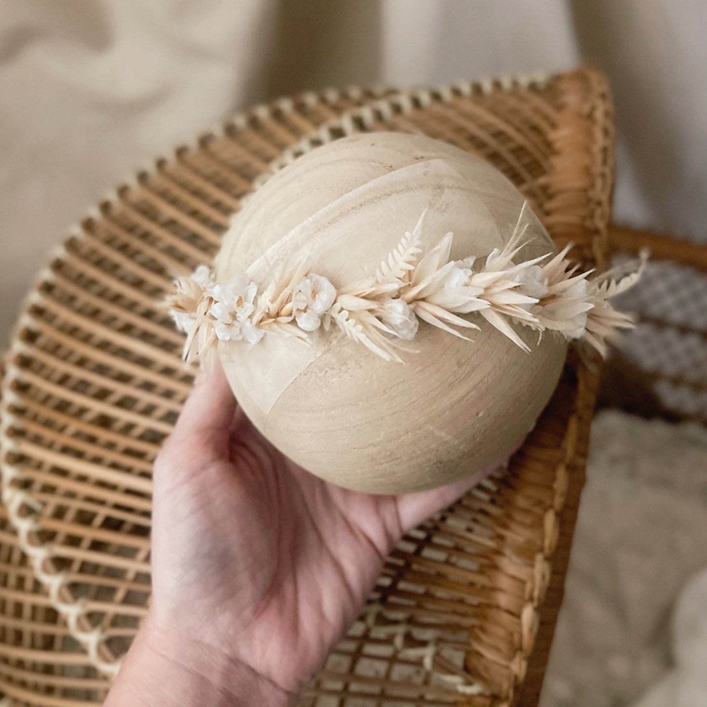 Hand holding a wooden sphere adorned with a delicate dried floral crown in neutral ivory and beige tones.