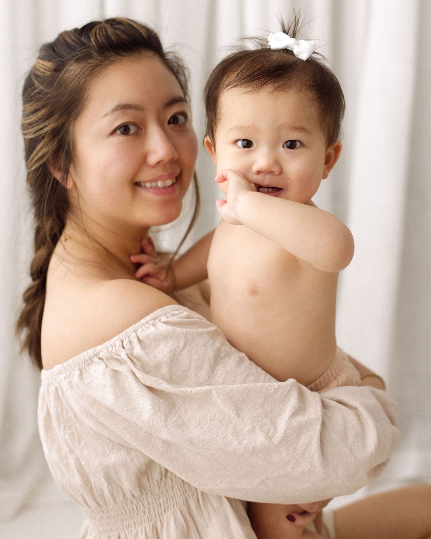 Smiling woman with a long side braid in a cream off-shoulder top holds a baby with a white bow against a soft white backdrop.