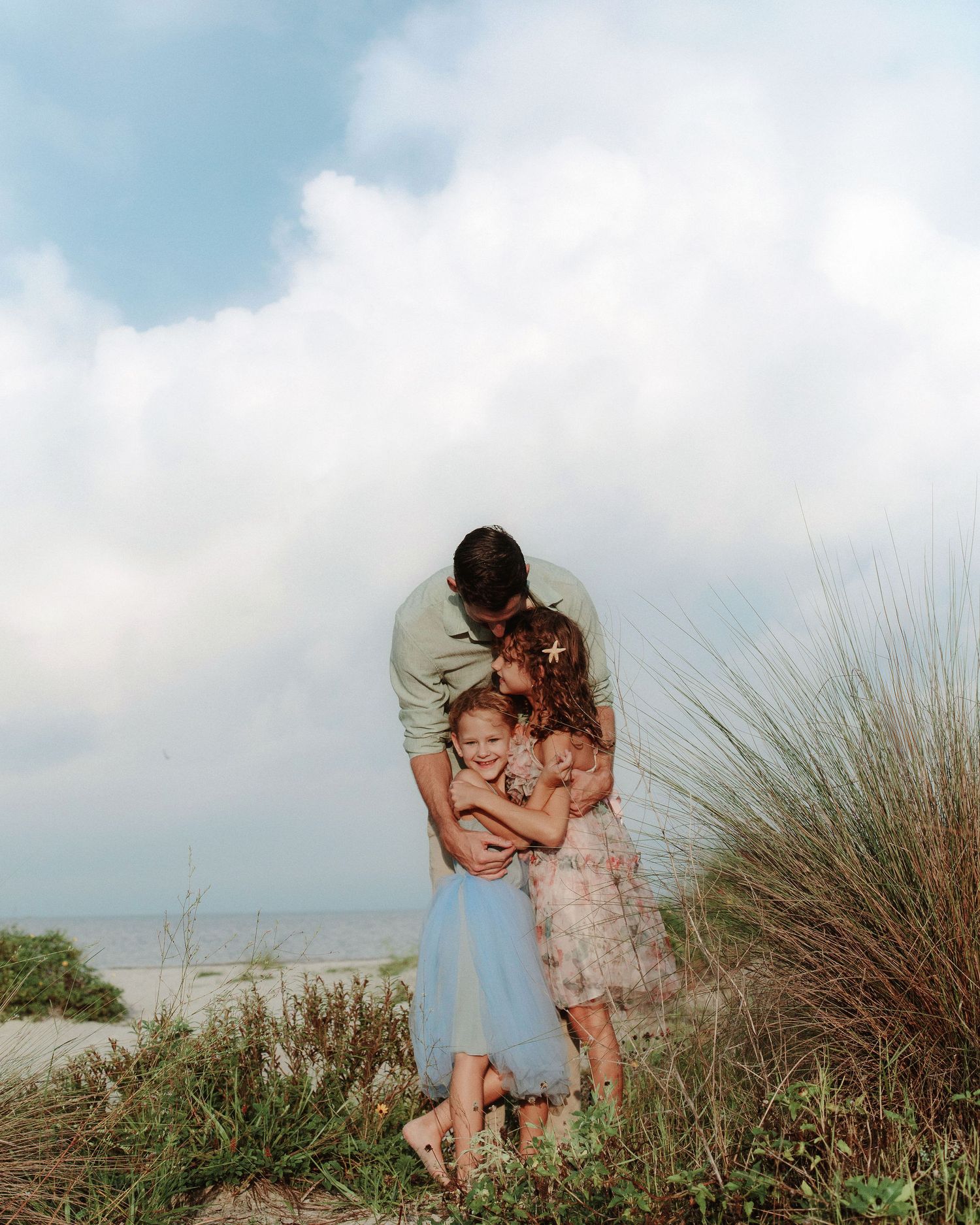 A man hugs two smiling young girls in dresses among beach grasses and sand dunes under a bright, cloudy sky near the ocean.