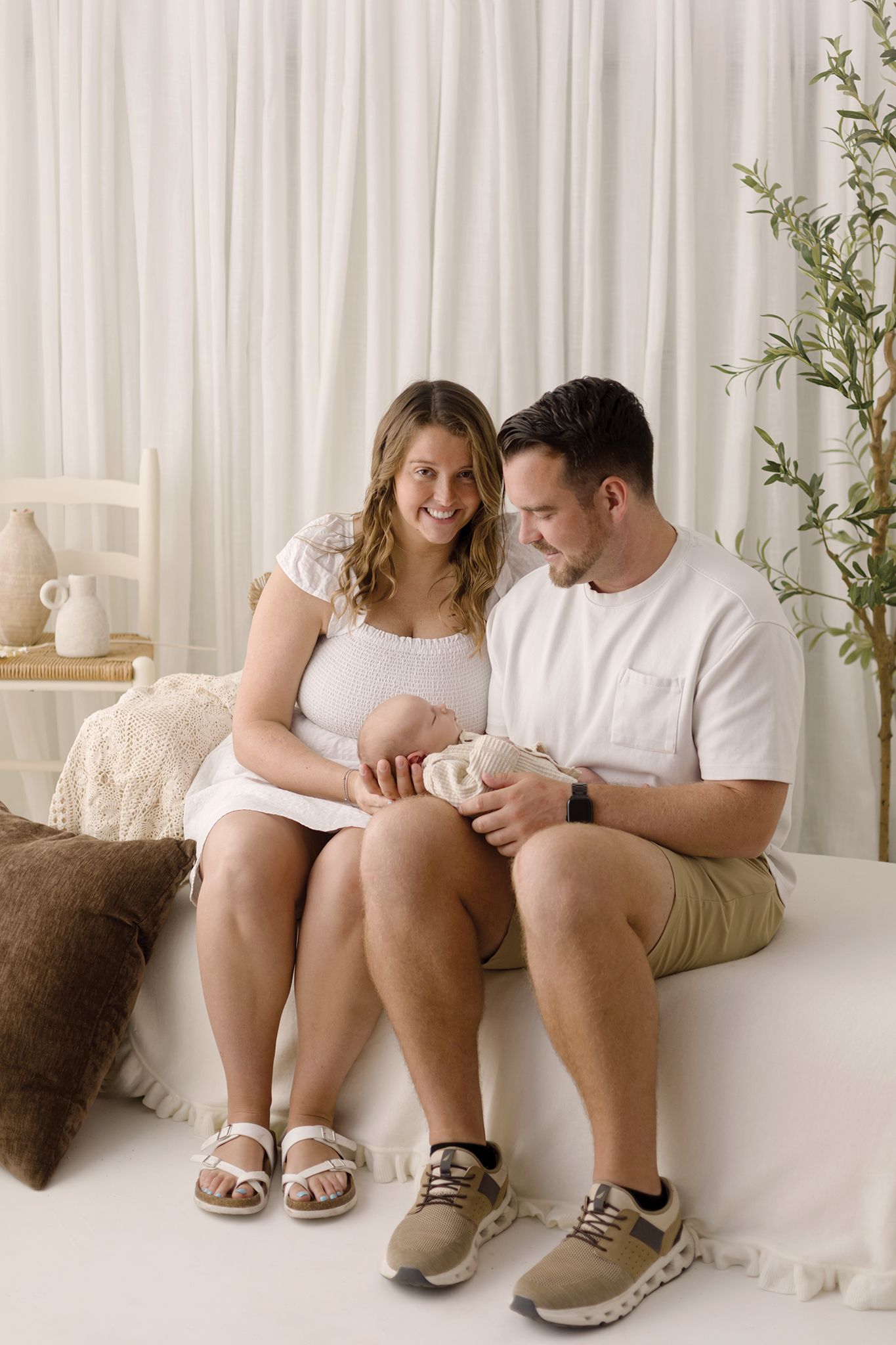 A happy couple sits together on a bed, lovingly cradling their newborn baby in a bright, neutral-toned studio setting.