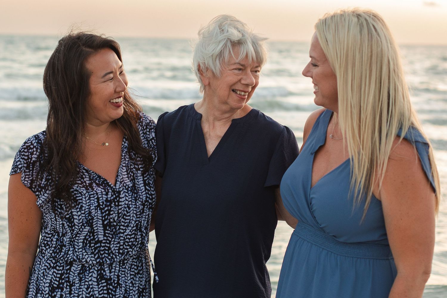 Three women smiling and laughing together on Redington beach at sunset, dressed in blue outfits.