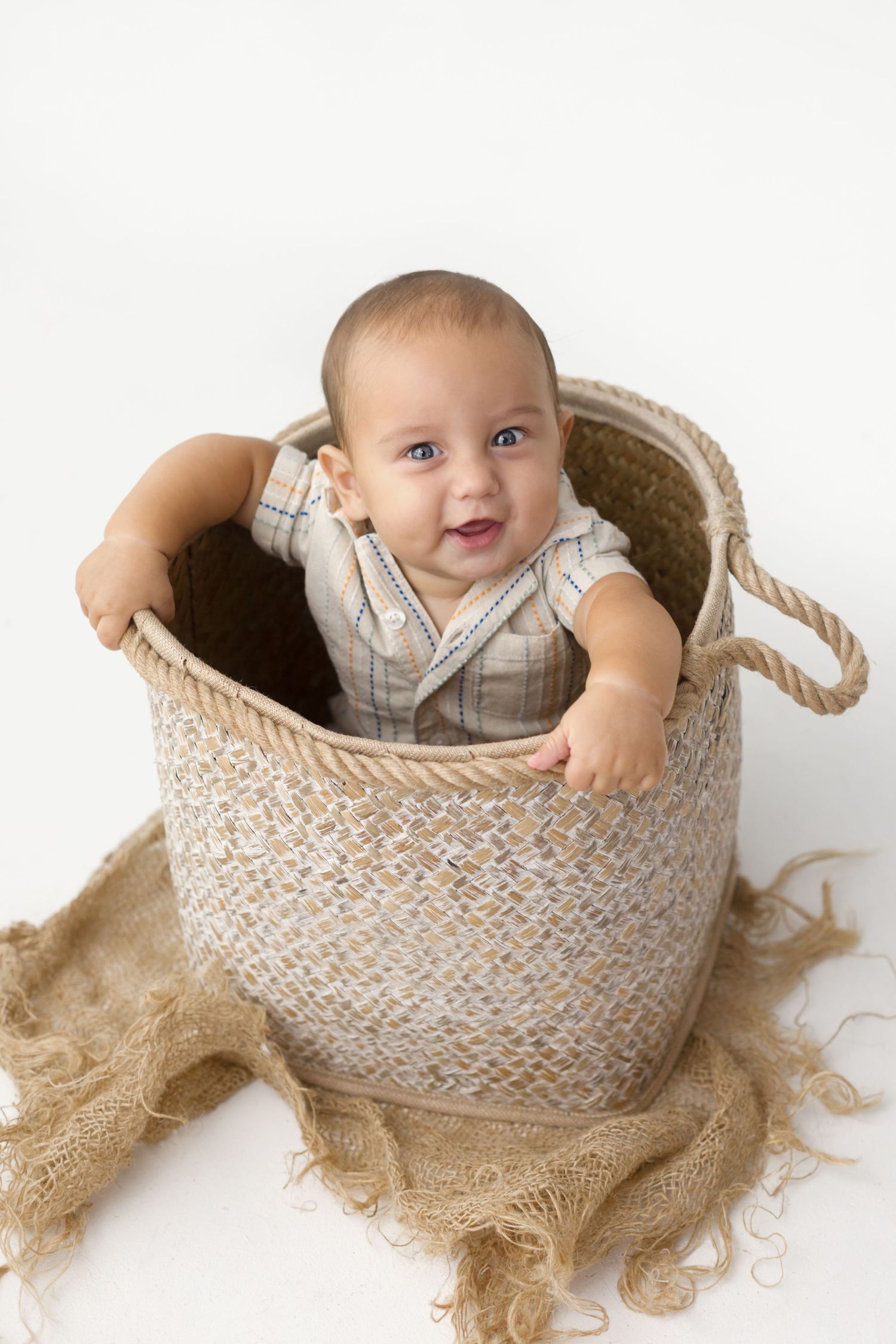 Smiling baby in a woven basket wearing a neutral romper, sitting on a white background with burlap fabric.
