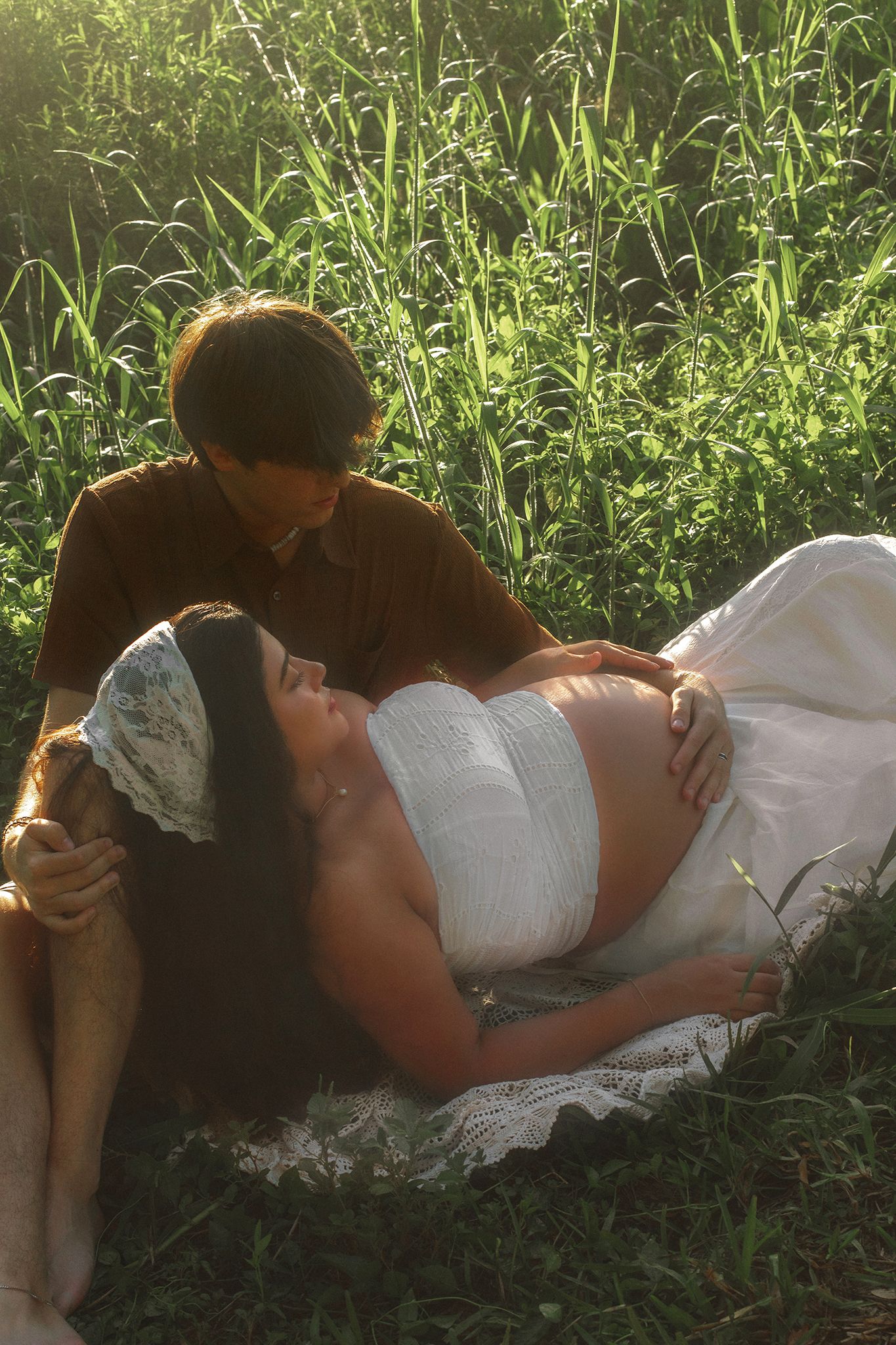 Pregnant woman in white dress lying in grass while partner in brown shirt gazes lovingly at her baby bump outdoors.