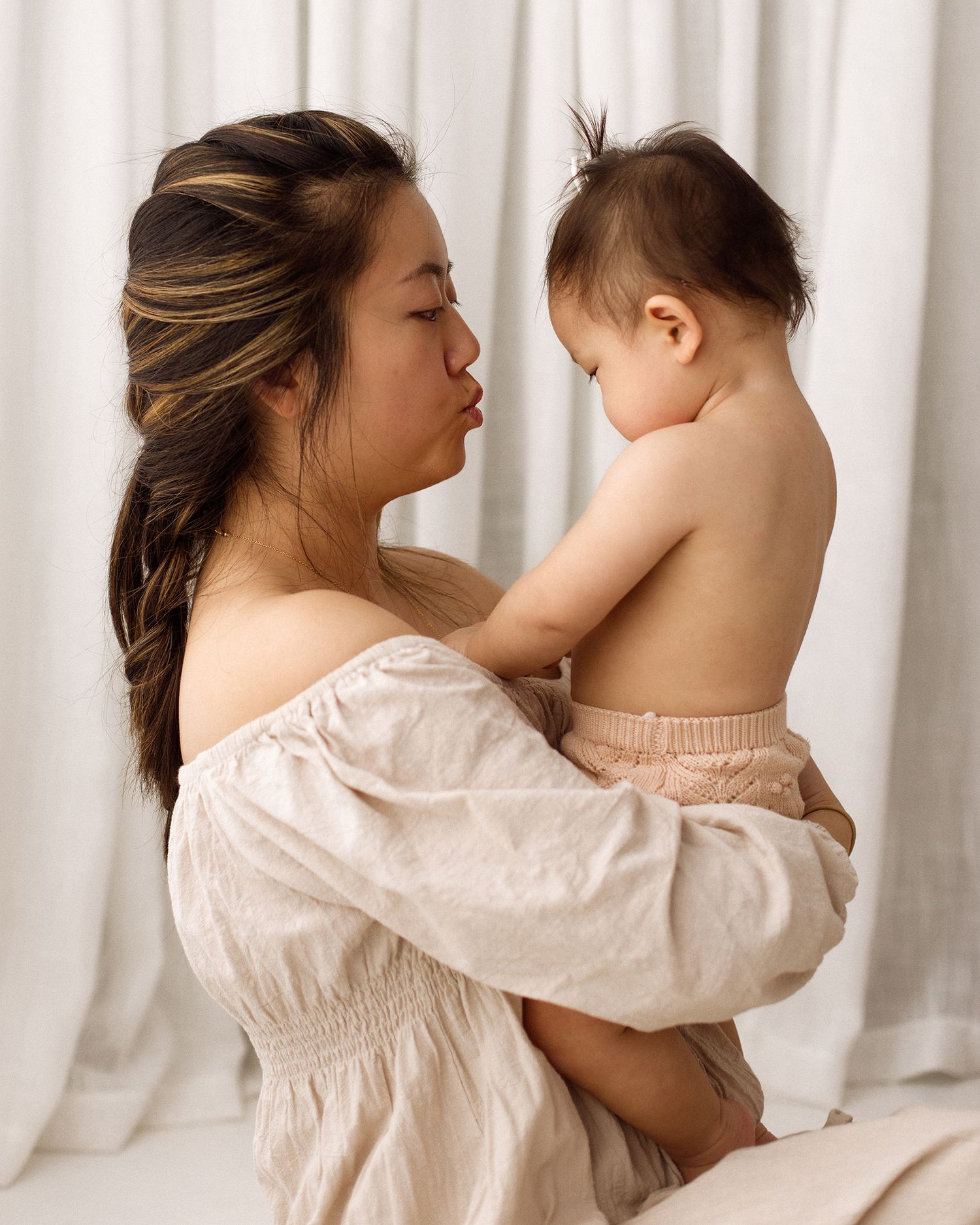 A woman in a cream off-shoulder blouse with a side braid holds a shirtless toddler and puckers her lips in front of soft white curtains.