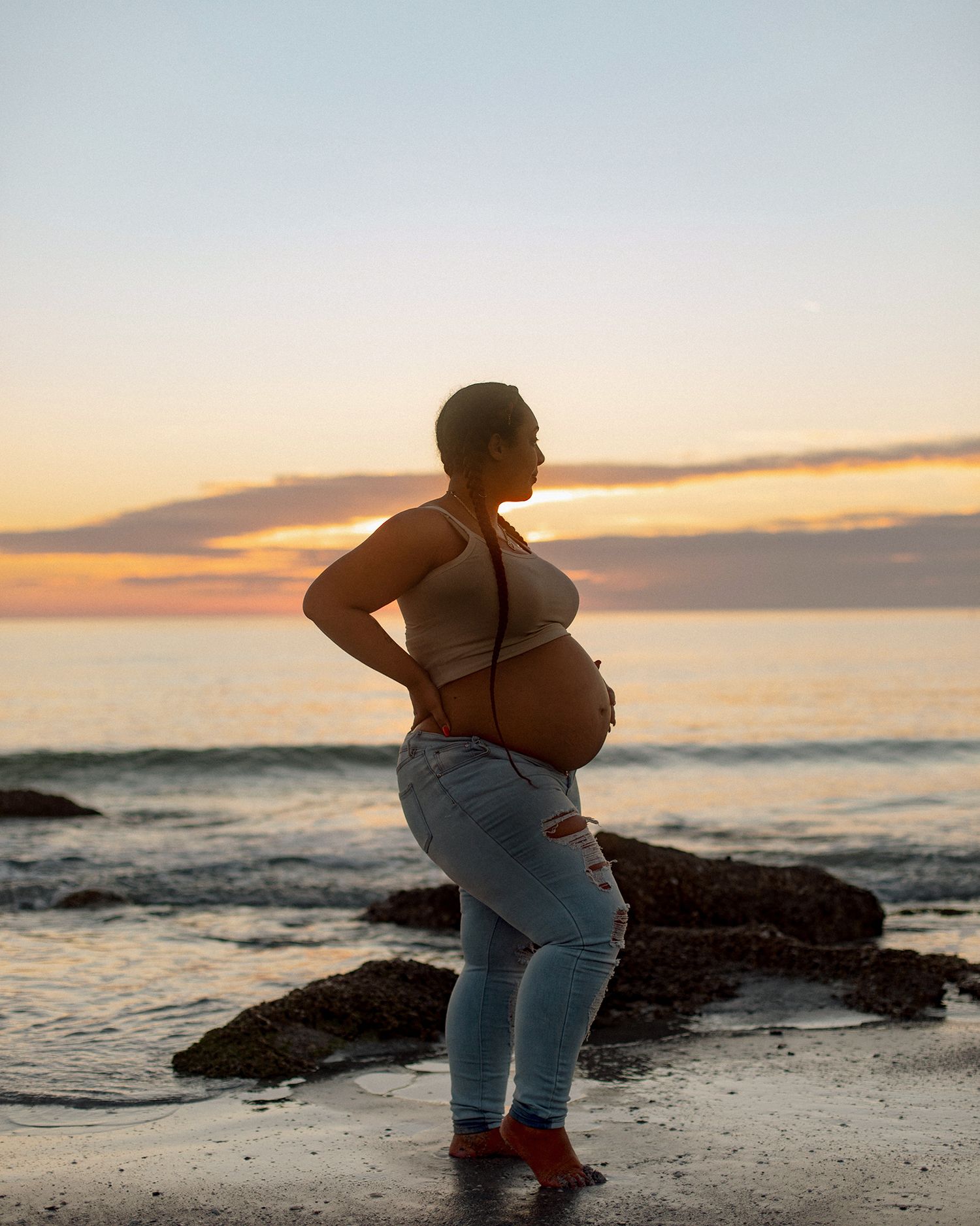 Pregnant woman in jeans stands on rocky beach at sunset, cradling her baby bump while gazing at the ocean.