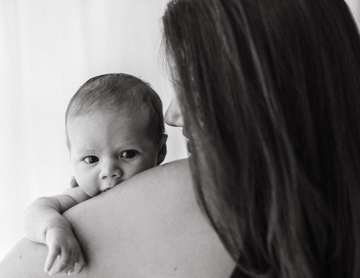 Black and white photo of a newborn baby resting on mother's shoulder, looking curiously at the camera.