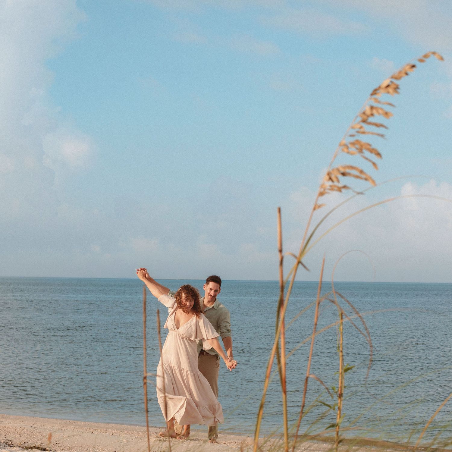 Couple in a flowing blush dress and casual outfit playfully dance on a sandy beach beside calm blue water and tall sea oats.