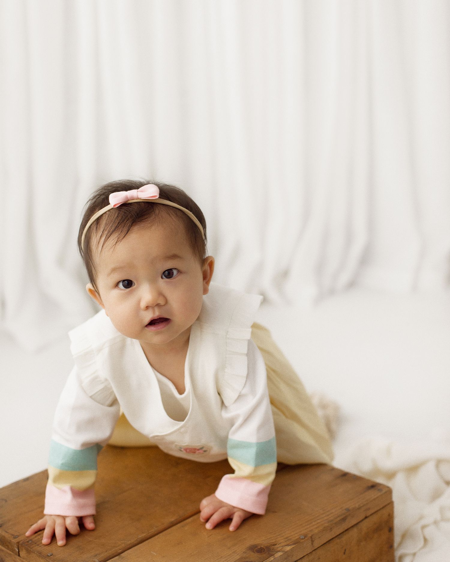 Baby girl in a pink bow headband and pastel striped outfit crawling on a rustic wooden crate against a white draped backdrop.