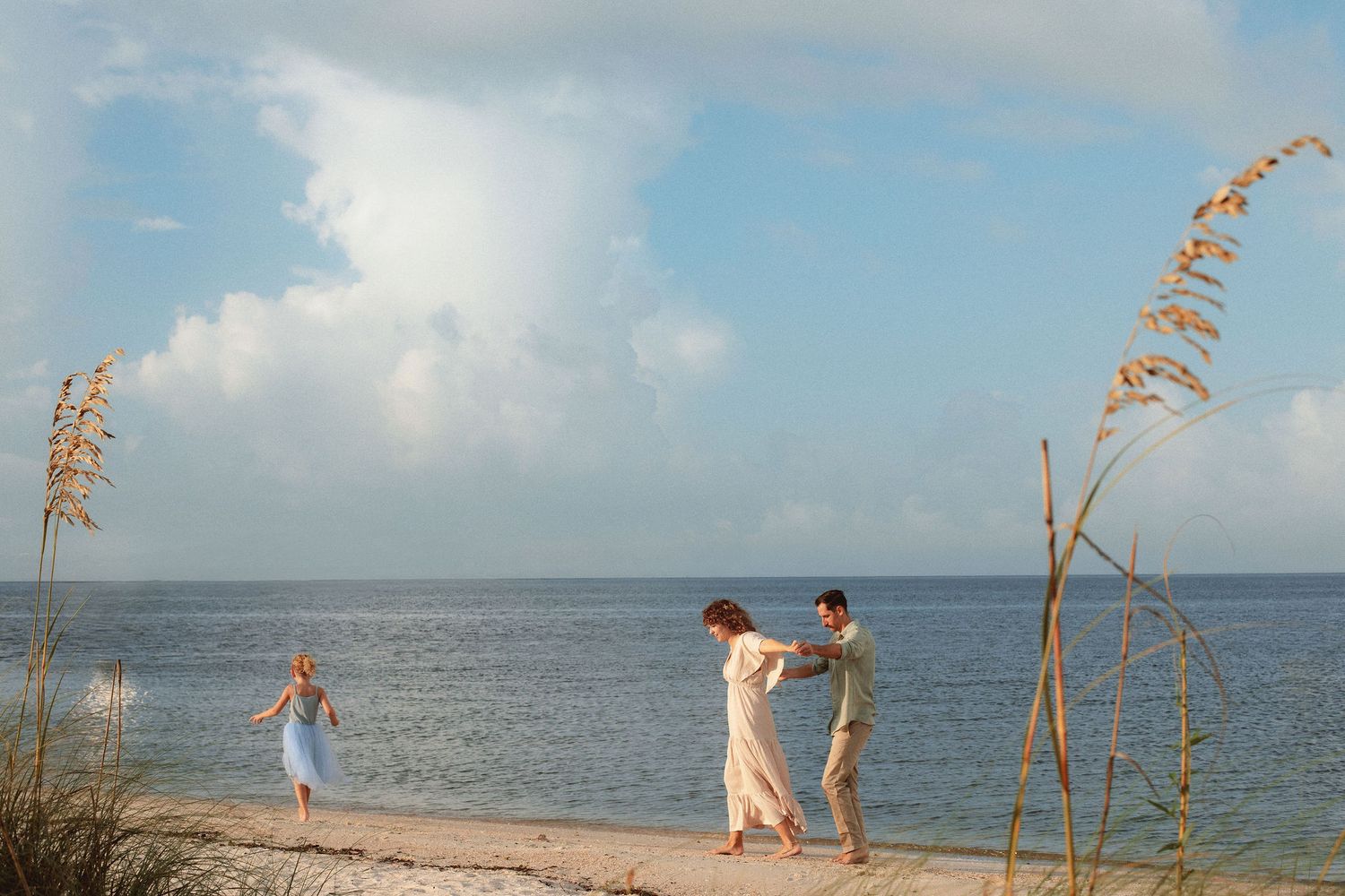 Couple in light summer clothing dance hand in hand on a sandy beach while a child plays nearby under a bright blue sky with puffy clouds.