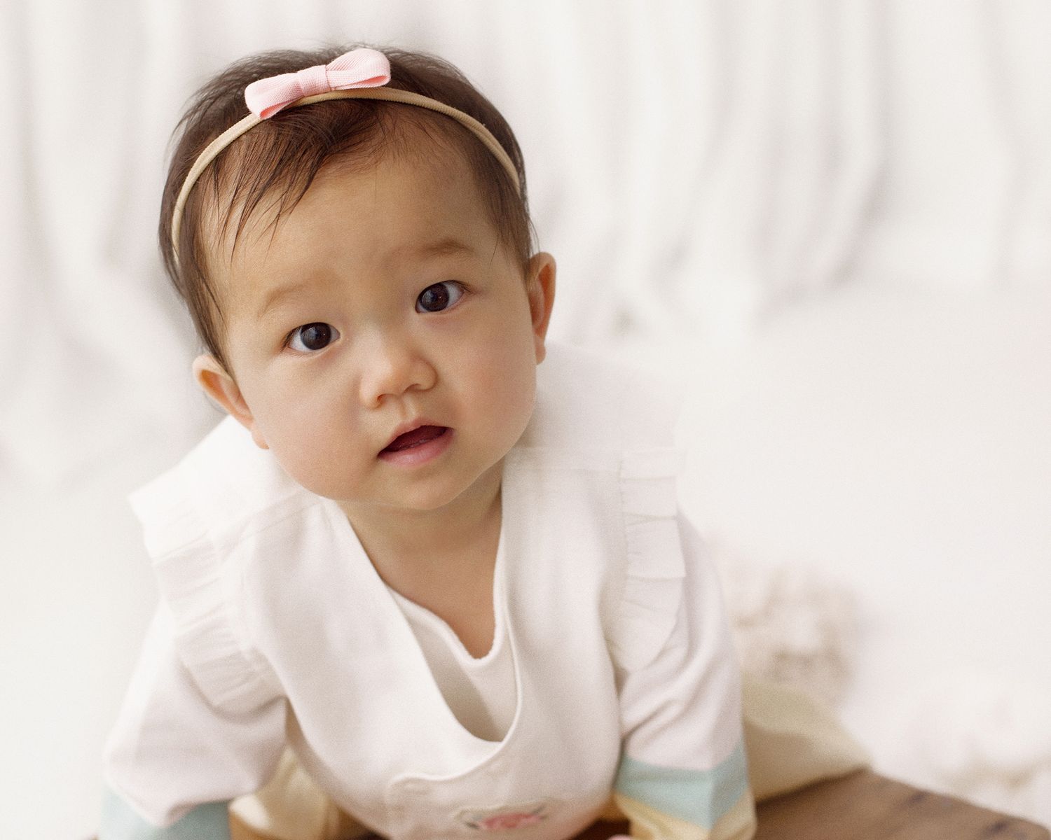 Baby girl in a white ruffled outfit and pink bow headband sits on a wooden floor against a soft white fabric backdrop.