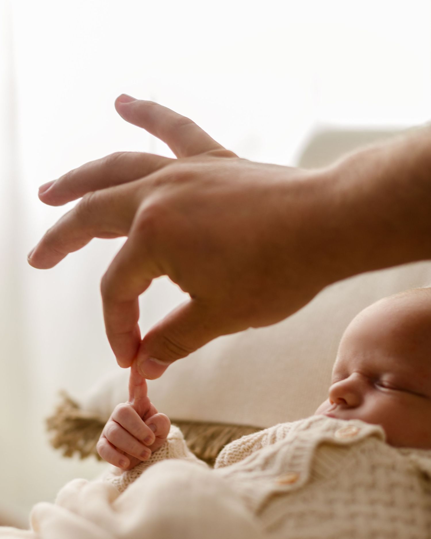 Close-up of a newborn baby grasping an adult finger while sleeping in a cream knit outfit.