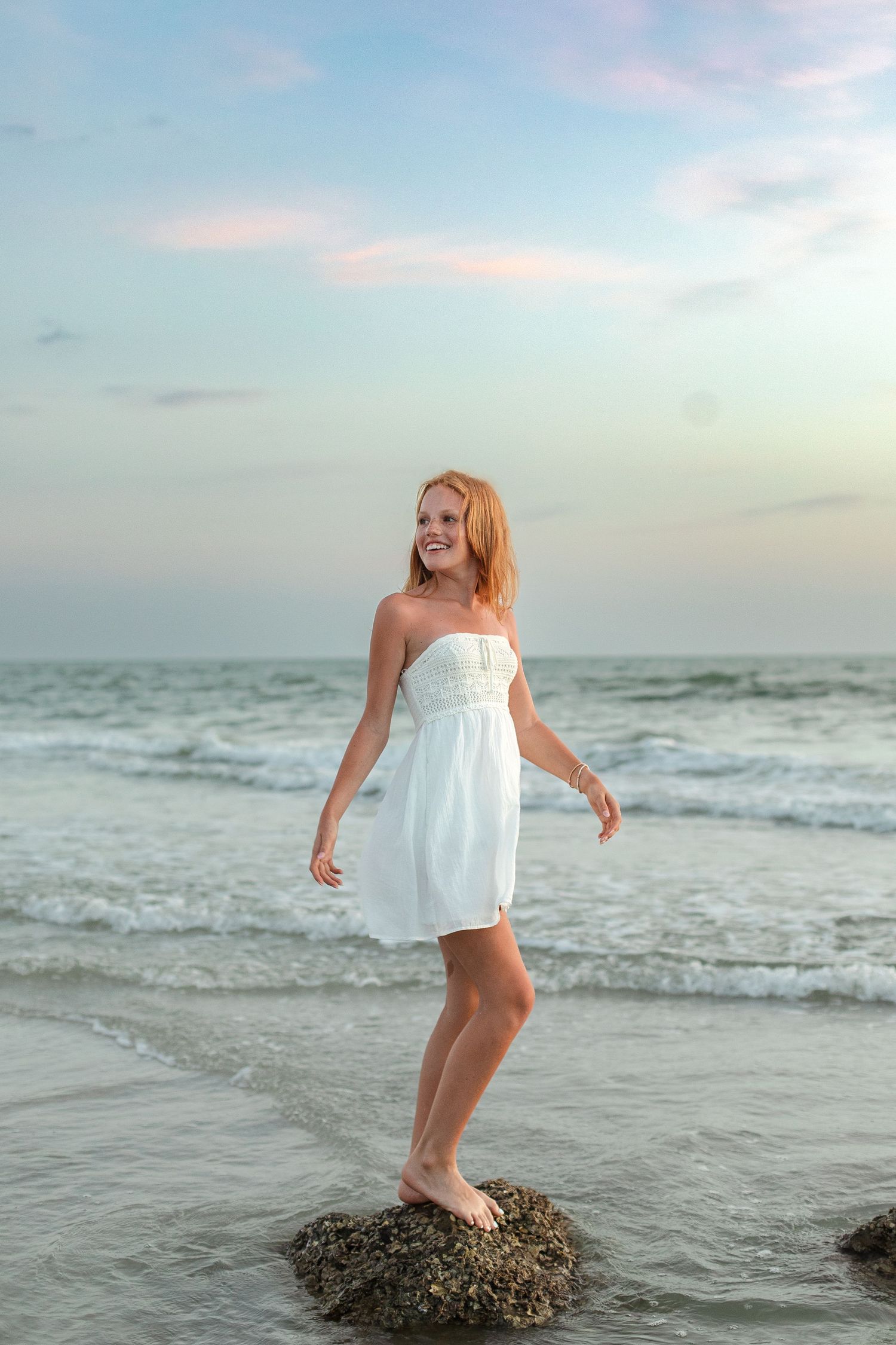 Young woman in white strapless dress standing on rocks at beach during sunset, smiling joyfully.