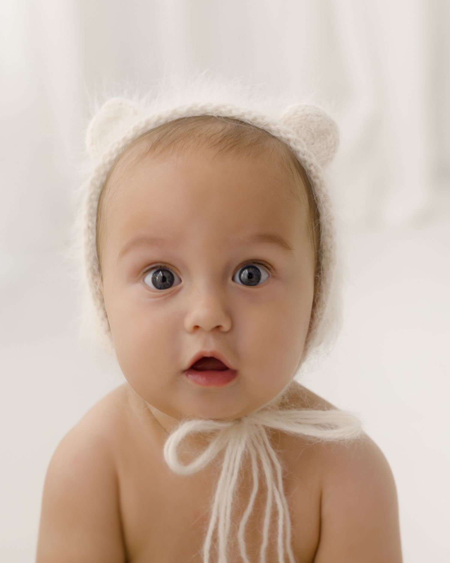 Adorable baby wearing a white fuzzy bear ear bonnet hat with tie strings, looking curiously at the camera.