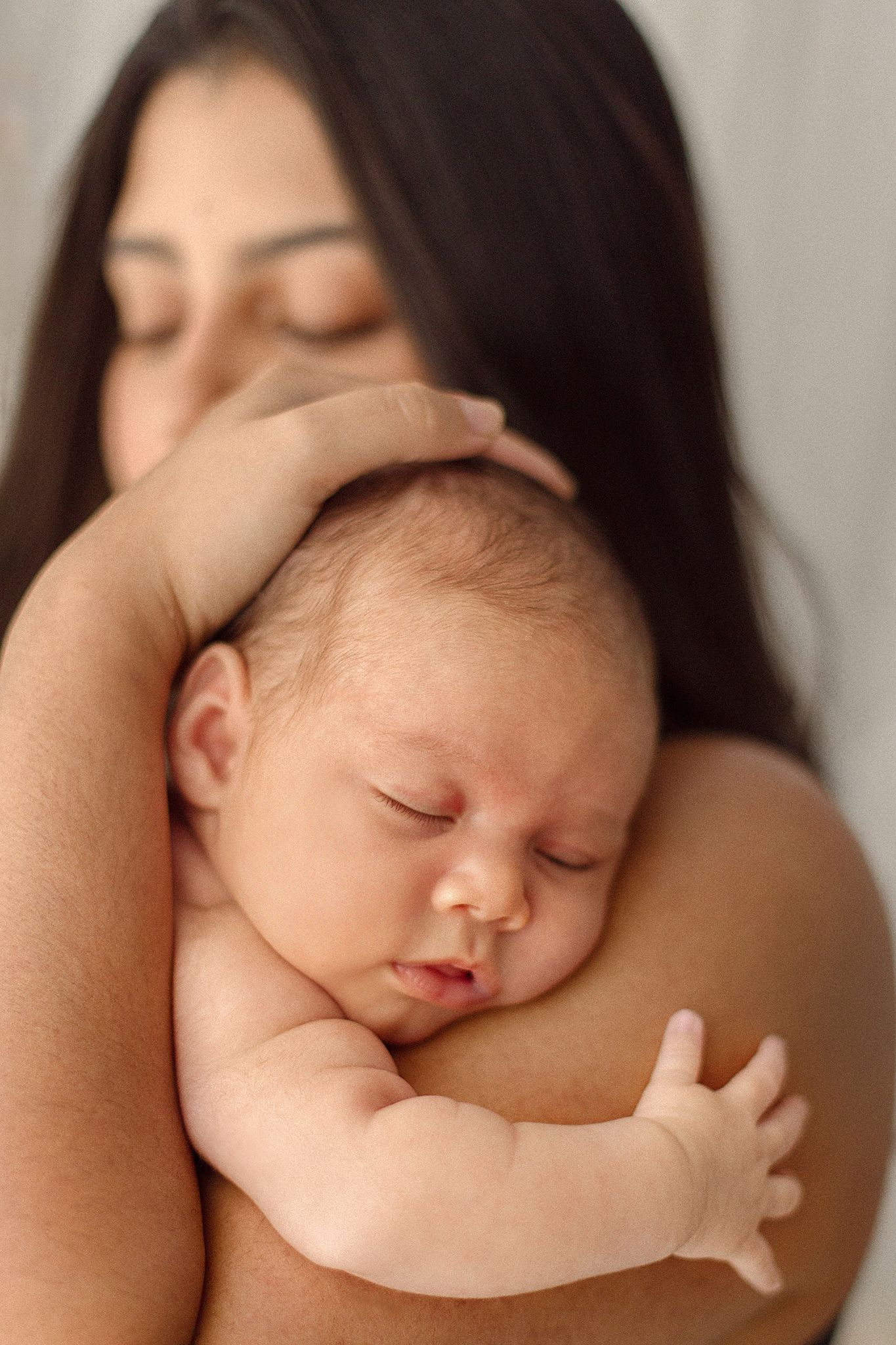 A mother with dark hair gently holds her sleeping newborn baby close to her chest in a tender embrace.