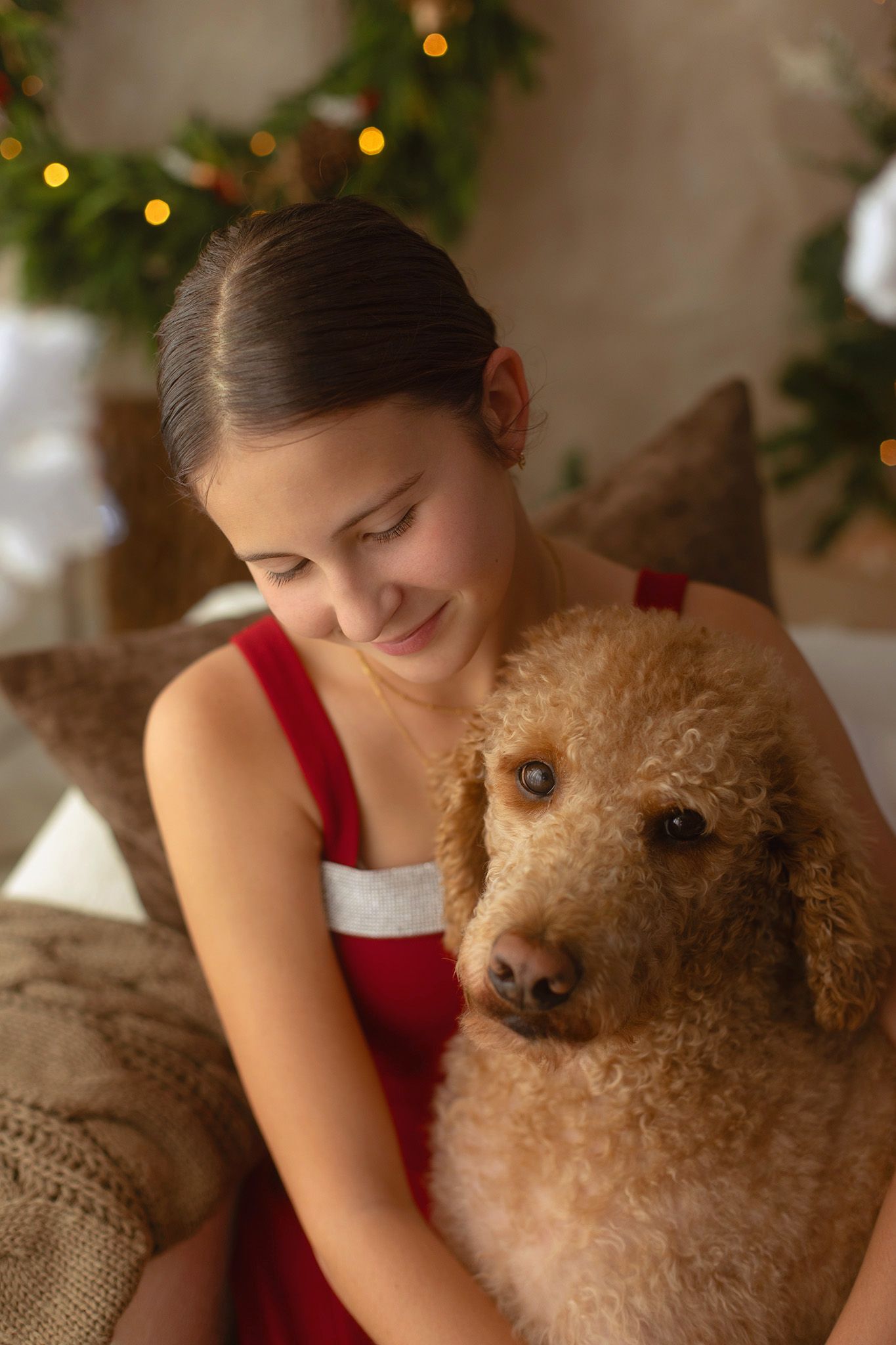 Young girl in red dress smiling while hugging a fluffy apricot poodle near a decorated Christmas tree.