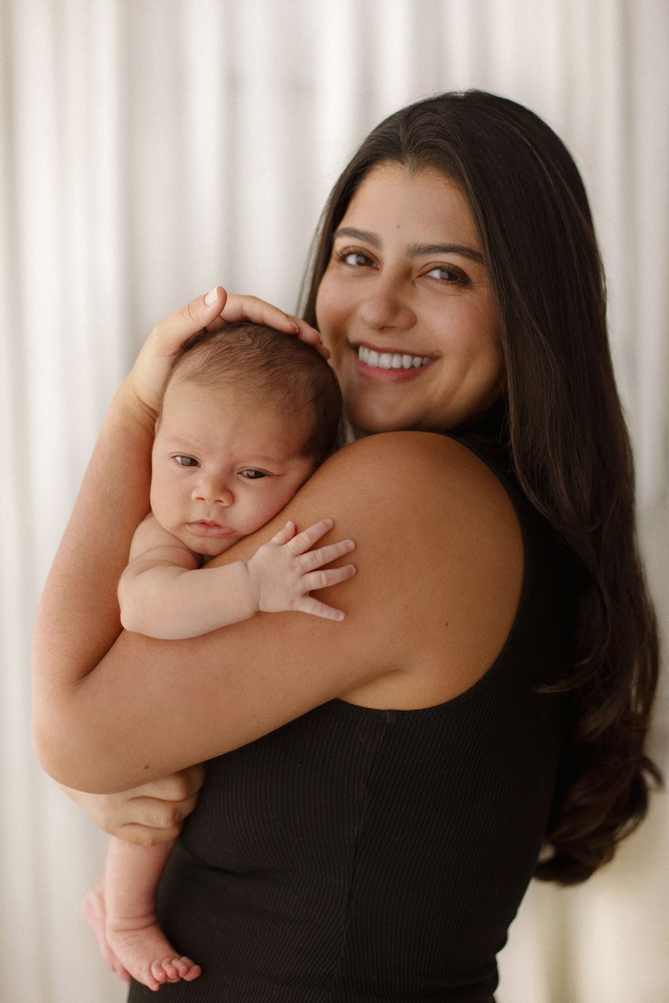 Smiling dark-haired woman in black top holding a newborn baby against white curtain background.