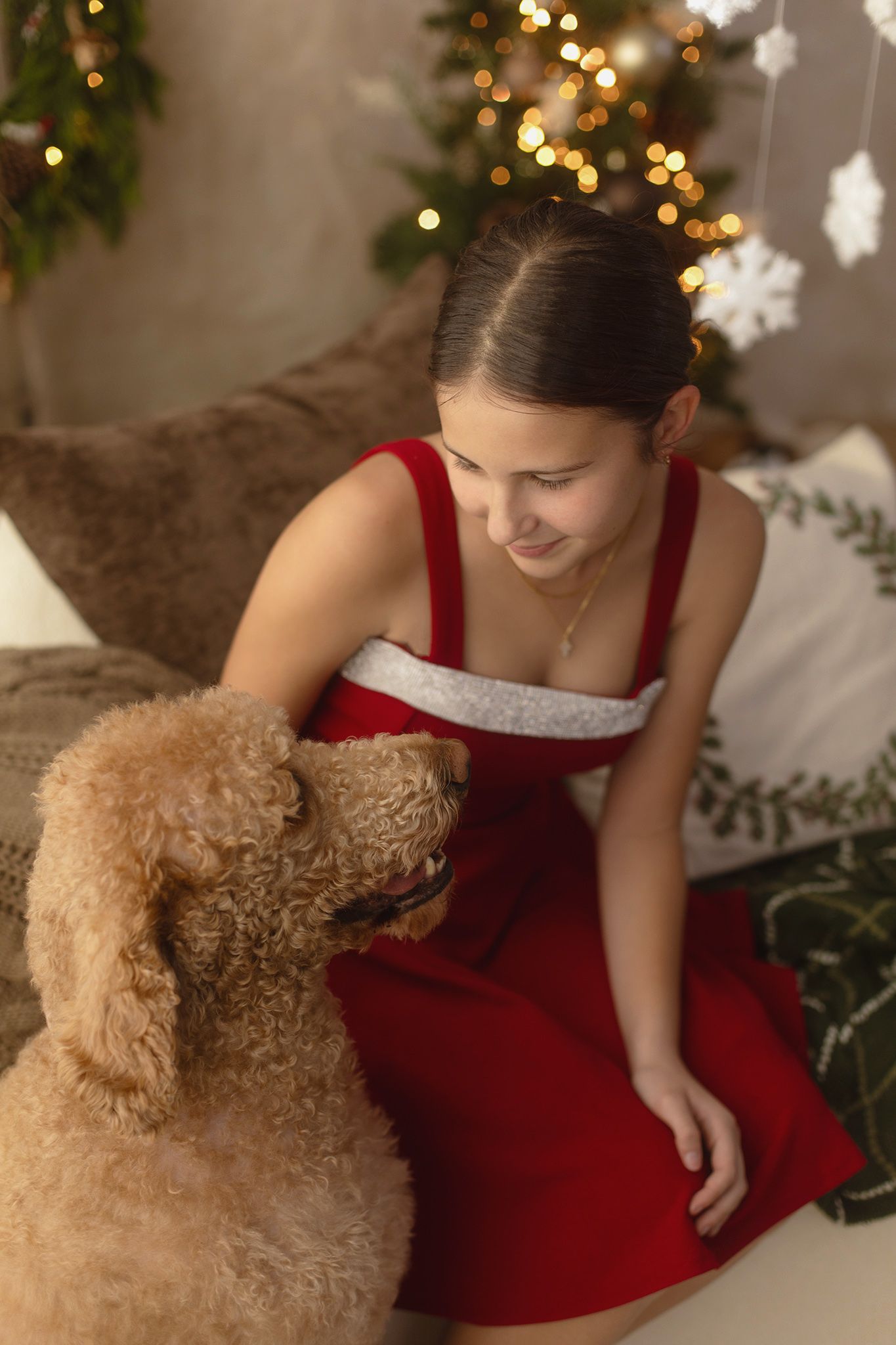 Young woman in red dress smiling at golden doodle dog wearing Santa hat near Christmas tree.