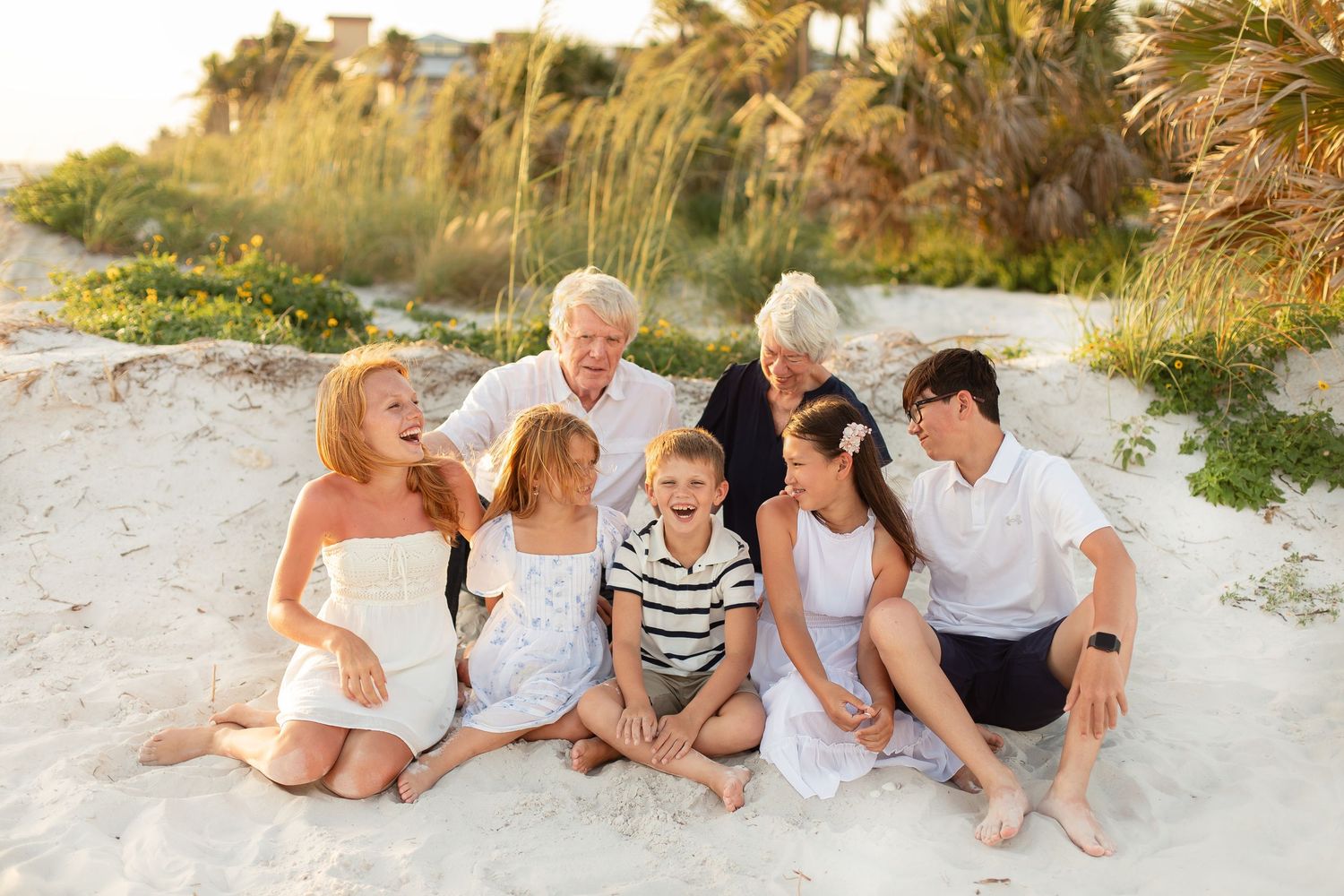 Multi-generational family smiling together on sandy beach at golden hour, dressed in white and blue outfits.