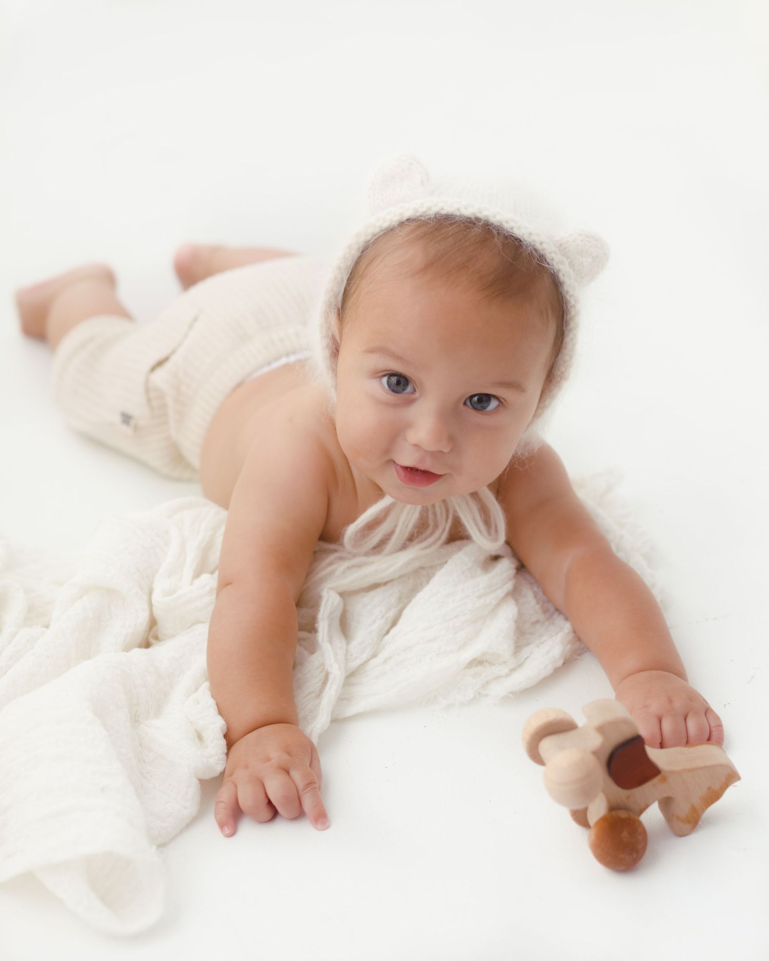 Adorable baby wearing a white bear bonnet lies on a soft white blanket, holding a wooden toy.