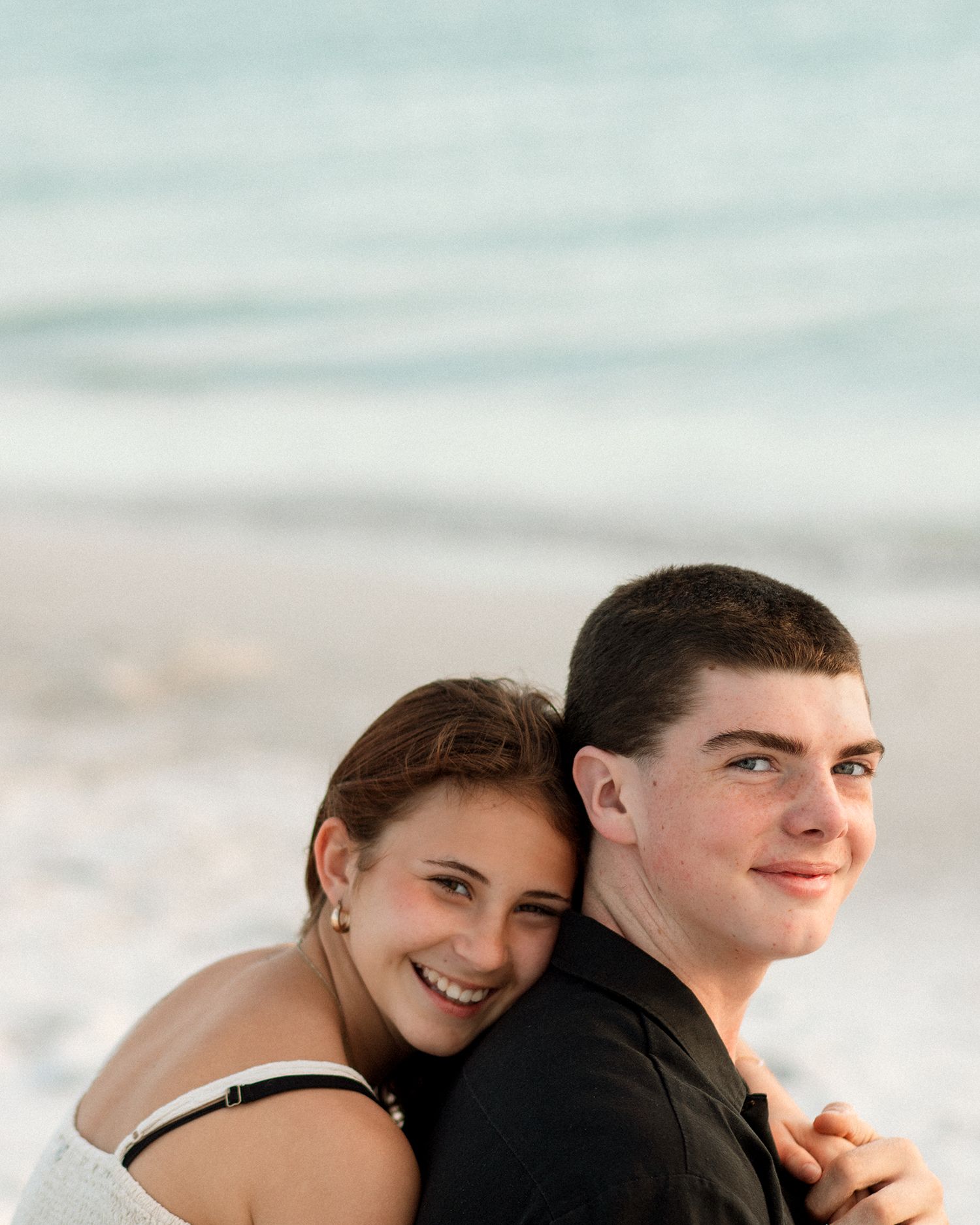 A young couple smiles together on a sandy beach, the woman hugging the man from behind near the ocean.
