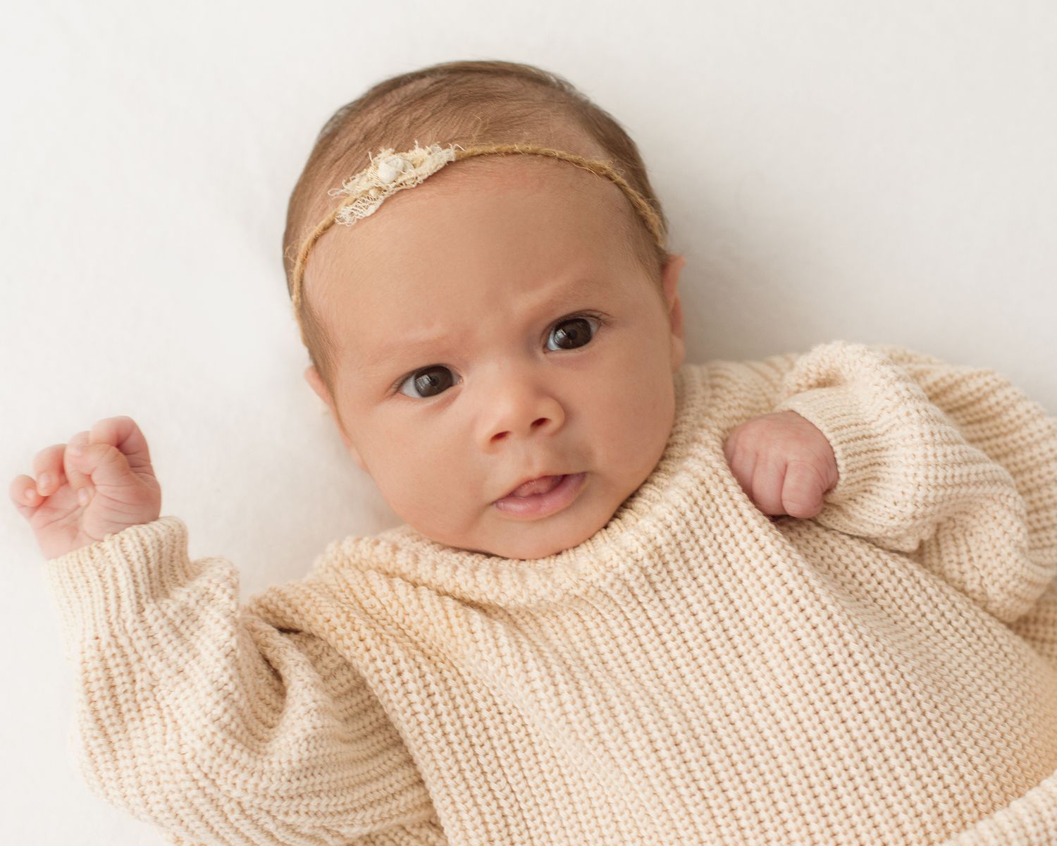 Newborn baby in cream knit sweater and gold headband lying on white background in St. Petersburg, Florida studio at one month old.