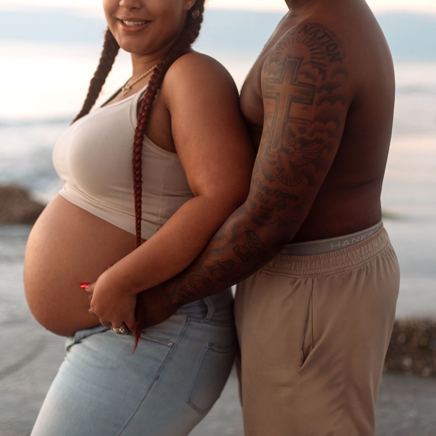 Pregnant woman in white crop top and jeans poses with tattooed partner at beach during golden hour.