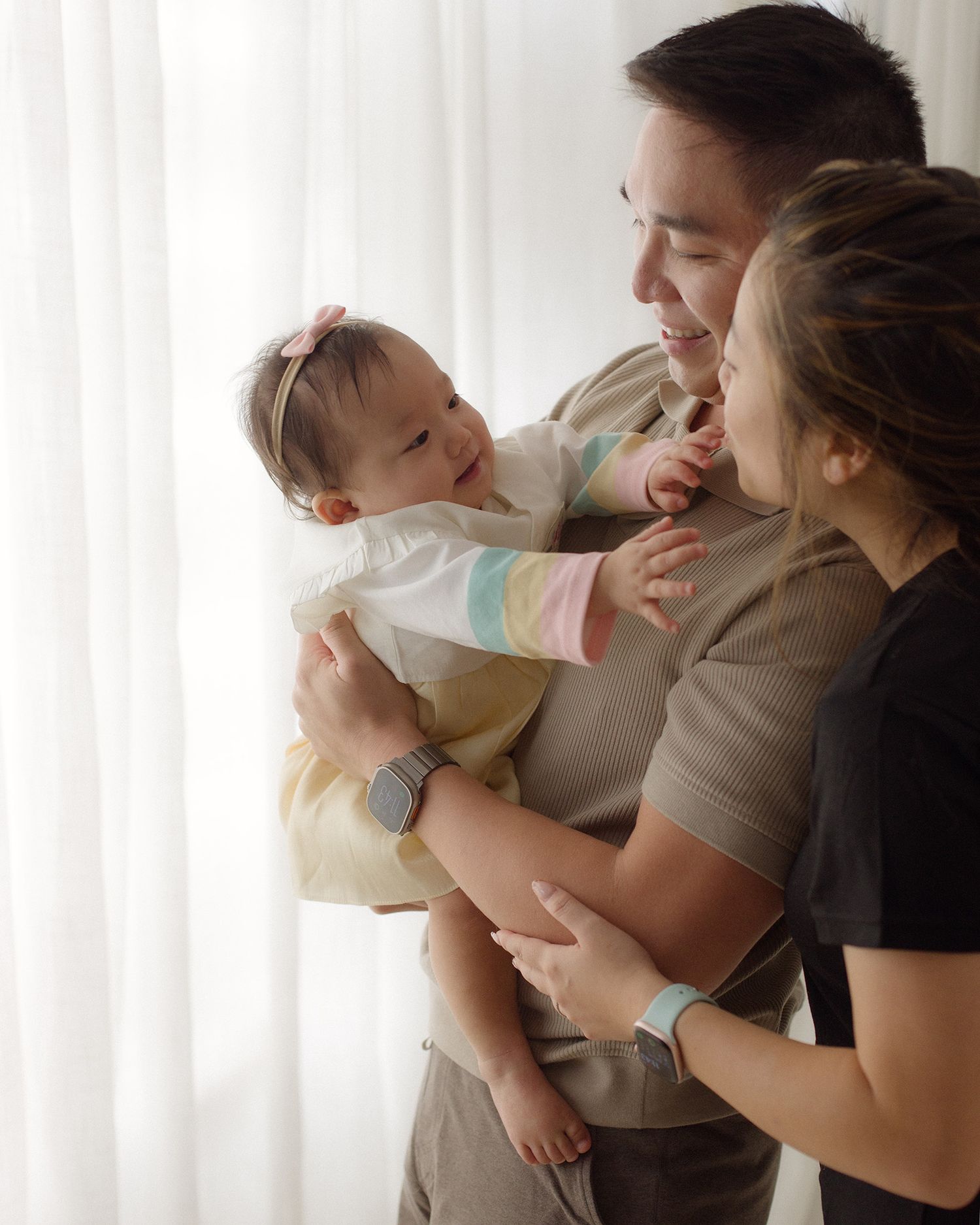 A smiling father and mother lovingly gaze at their baby girl, who wears a pink bow and rainbow-striped outfit, by a sunlit sheer curtain.
