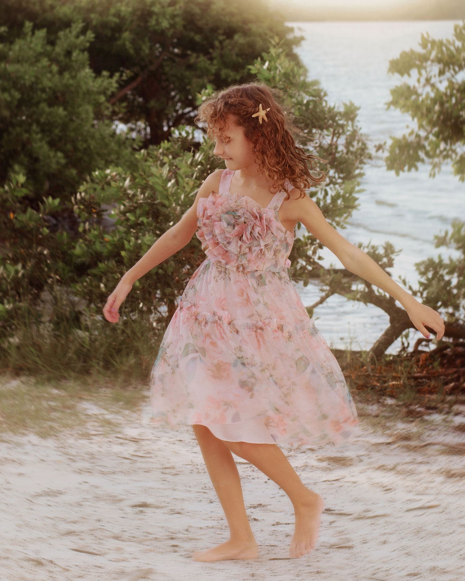 Young girl in a pink floral chiffon dress and star hair clip dances barefoot on a sandy beach near green trees and calm water.