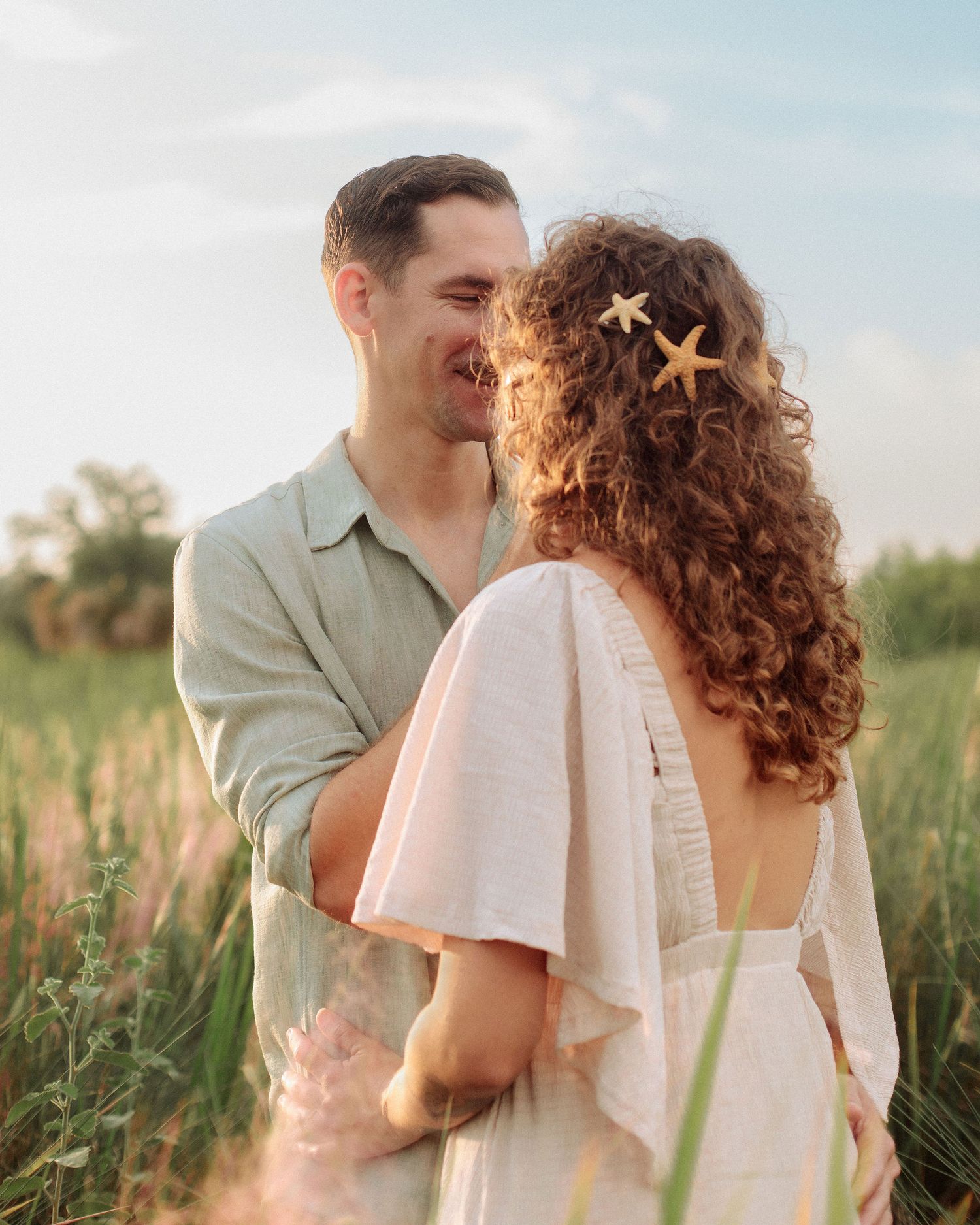 Smiling couple embracing in a sunny grassy field, the woman with curly hair adorned with starfish clips, in a romantic golden hour portrait.