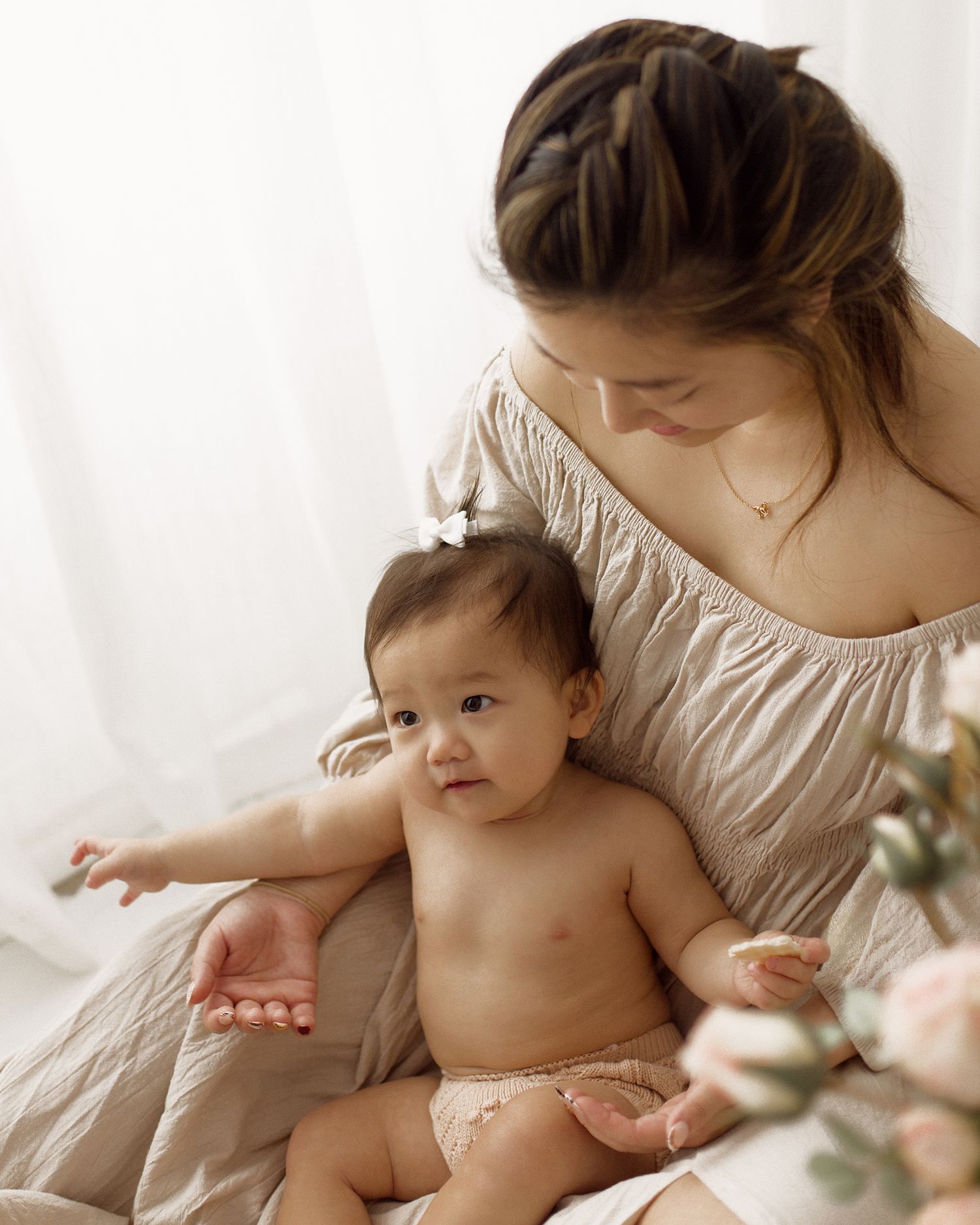 A woman with a braided updo in a beige off-shoulder dress gently holds a smiling baby in a bow clip, with soft flowers in the foreground.