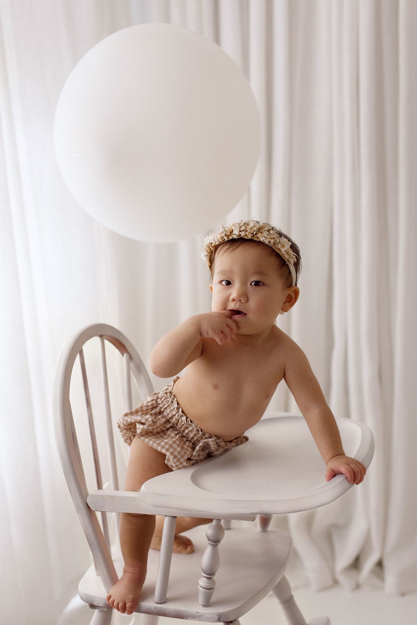 Baby in a gingham diaper cover and floral headband stands on a white wooden high chair beside a large white balloon.