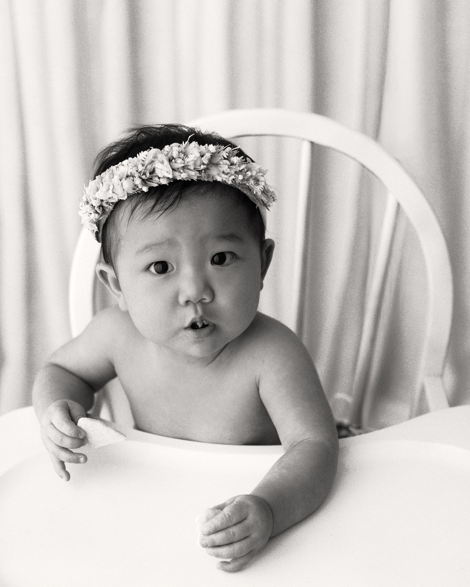 Black and white portrait of a baby in a white high chair wearing a floral flower crown headband, with hands resting on the tray.