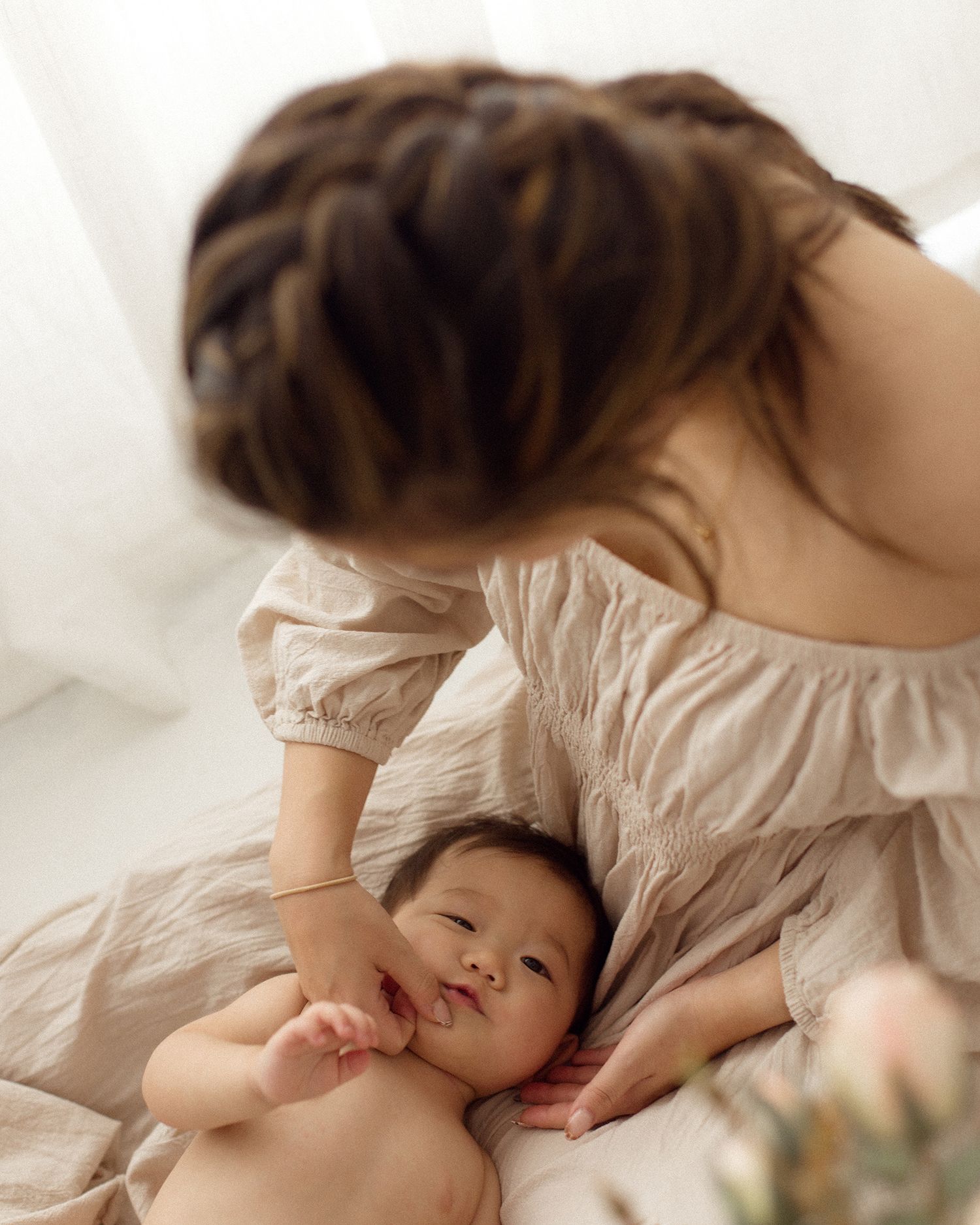 A mother with a braided hairstyle leans over a smiling baby lying on soft beige bedding in bright natural light.